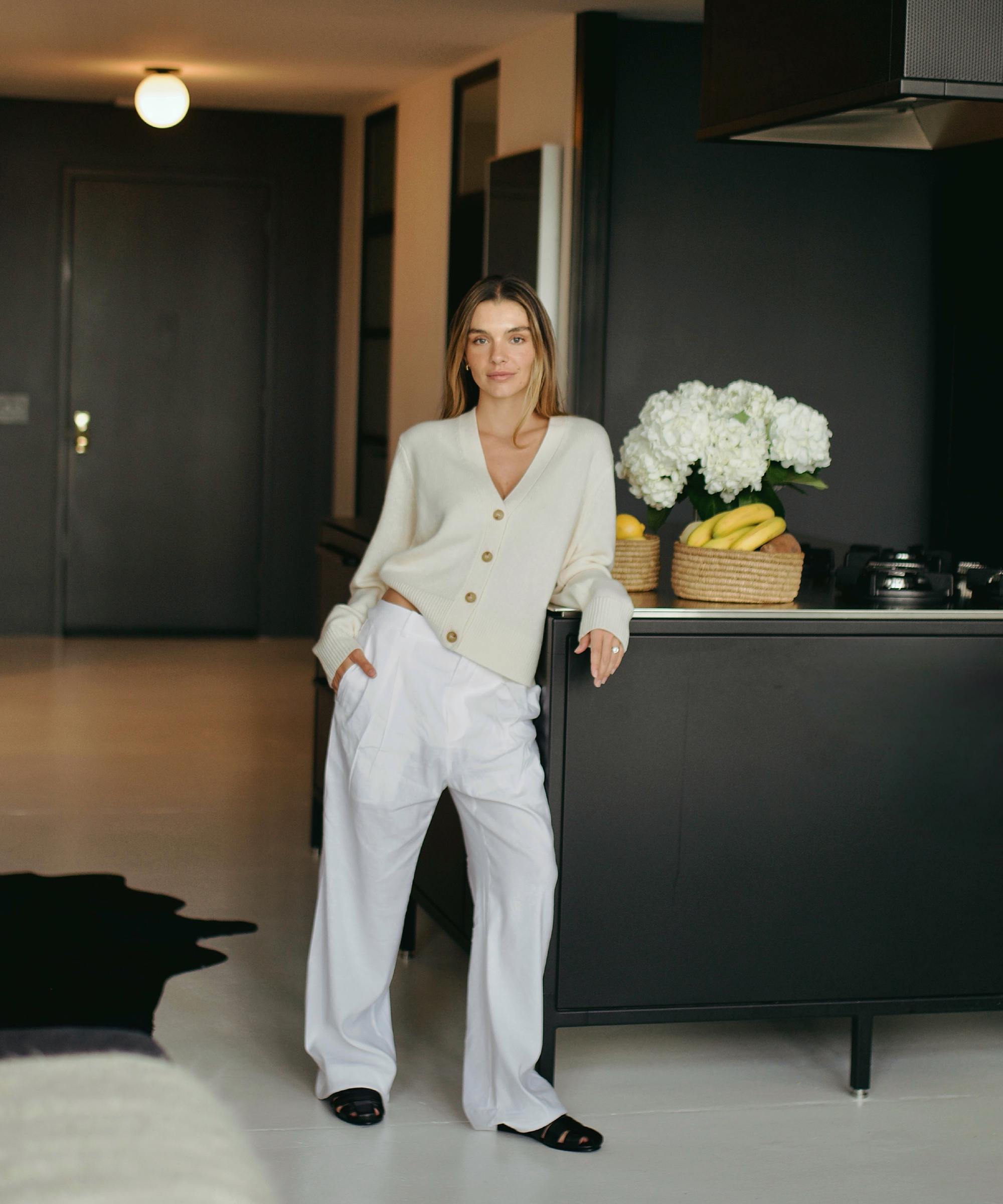 lauren burke in white trousers and an ivory cashmere cardigan standing in a black kitchen with black leather mules