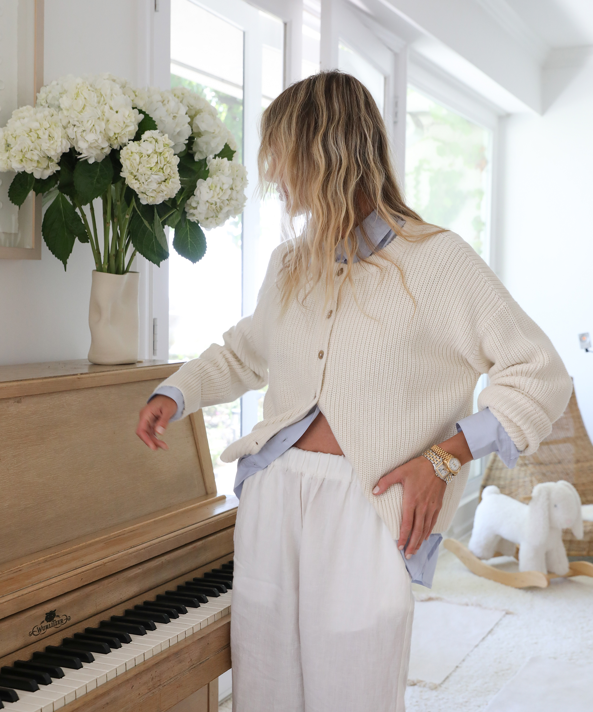 Cyd Morris, a woman with wavy blonde hair, wearing a cream cardigan and light blue shirt, stands beside a light wood piano topped with a vase of white flowers in a bright, cozy room.