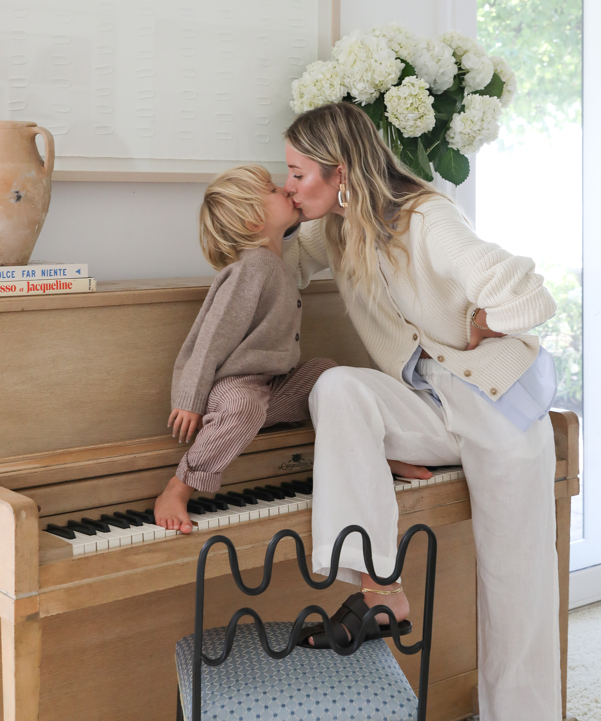 A woman and a young child share a kiss while the child sits barefoot on a piano keyboard. The woman, possibly Cyd Morris, leans in smiling, dressed in light clothing. A vase of white flowers and a jug decorate the piano.