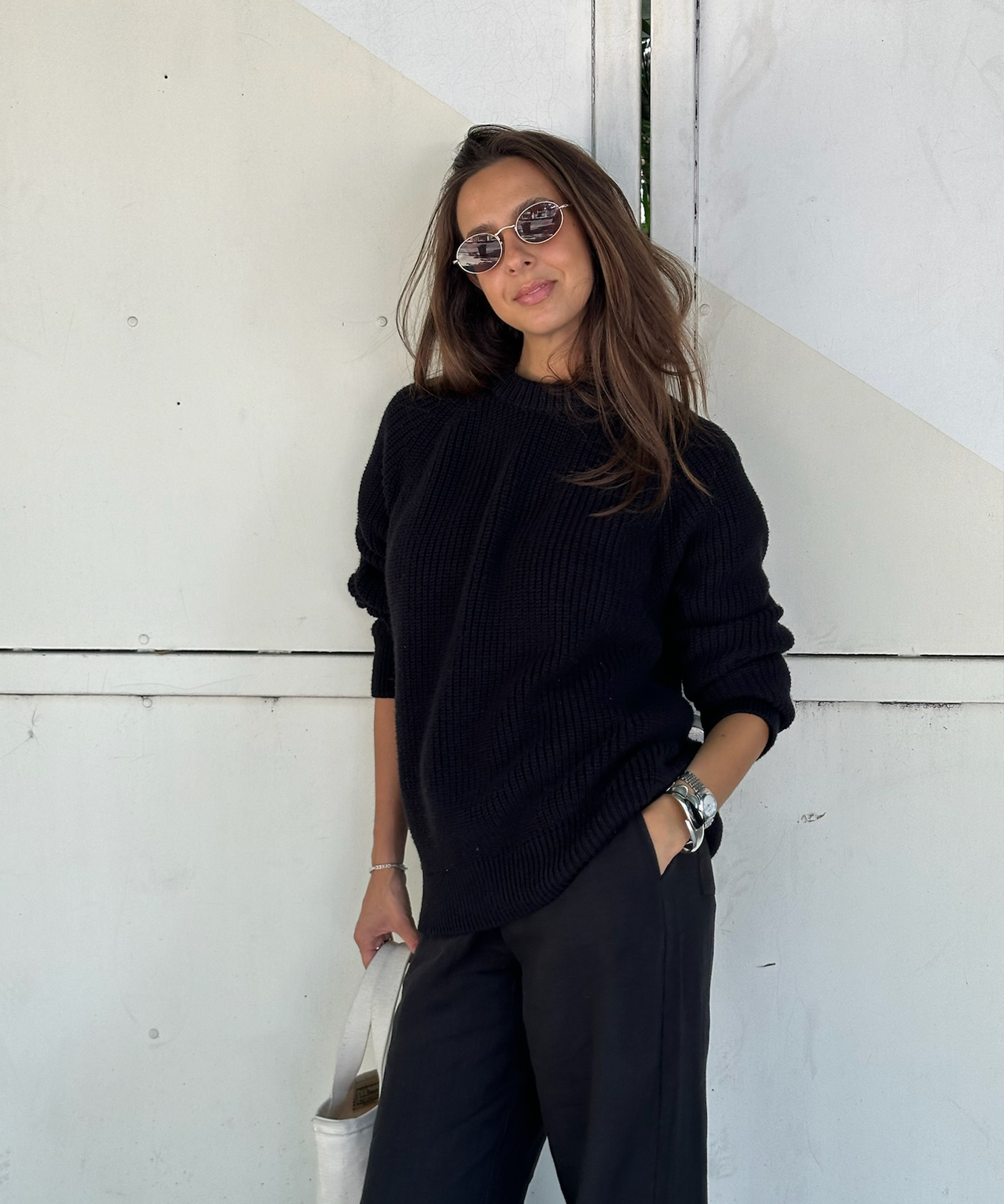 gabriella in an oversized cotton knit and black linen pants leaning against an ivory wall