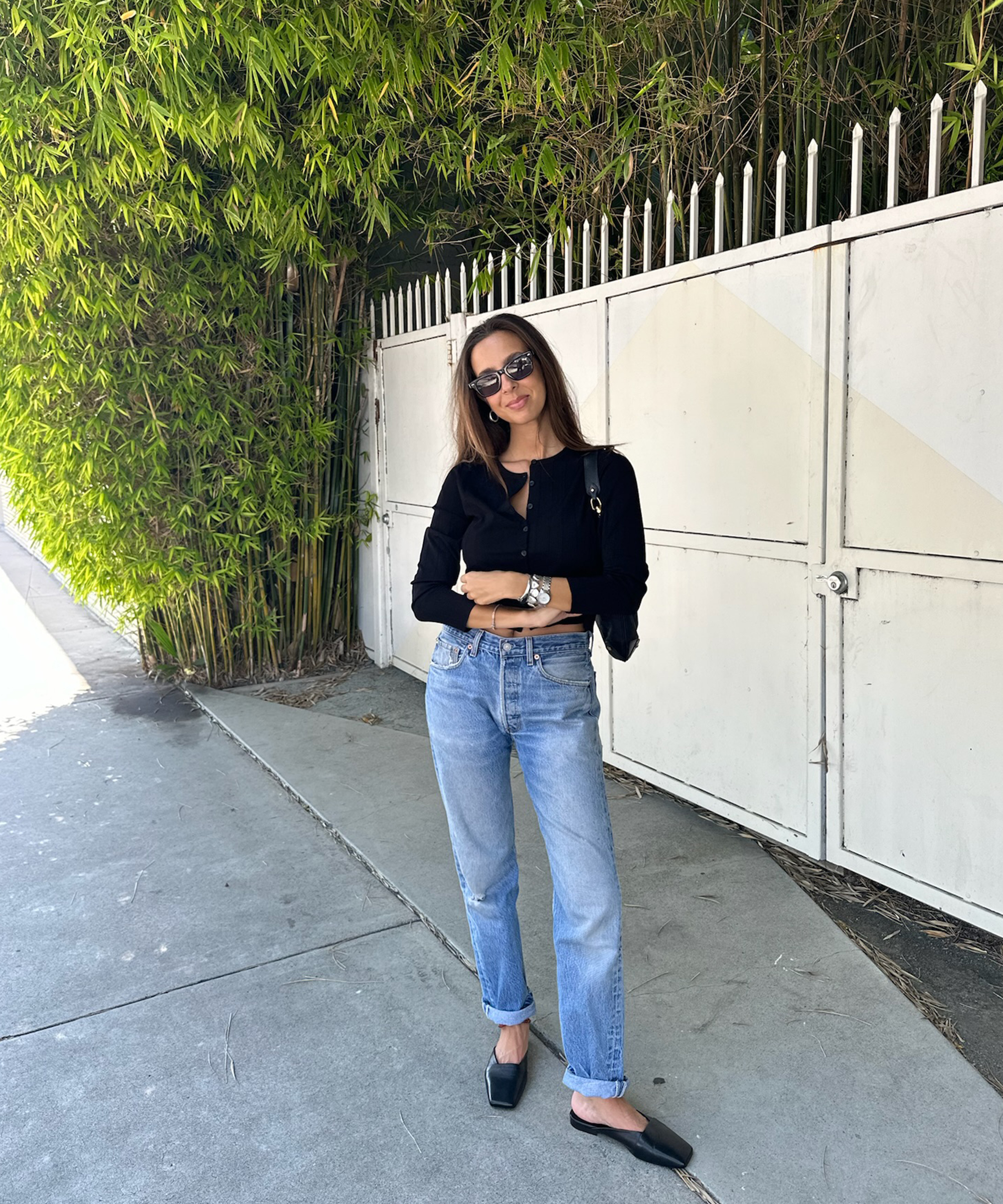 Gabriella Gofis stands on a sidewalk in front of a white fence and tall green bamboo, wearing sunglasses, a black long-sleeve top, light blue jeans, black flats, and carrying a small black bag.