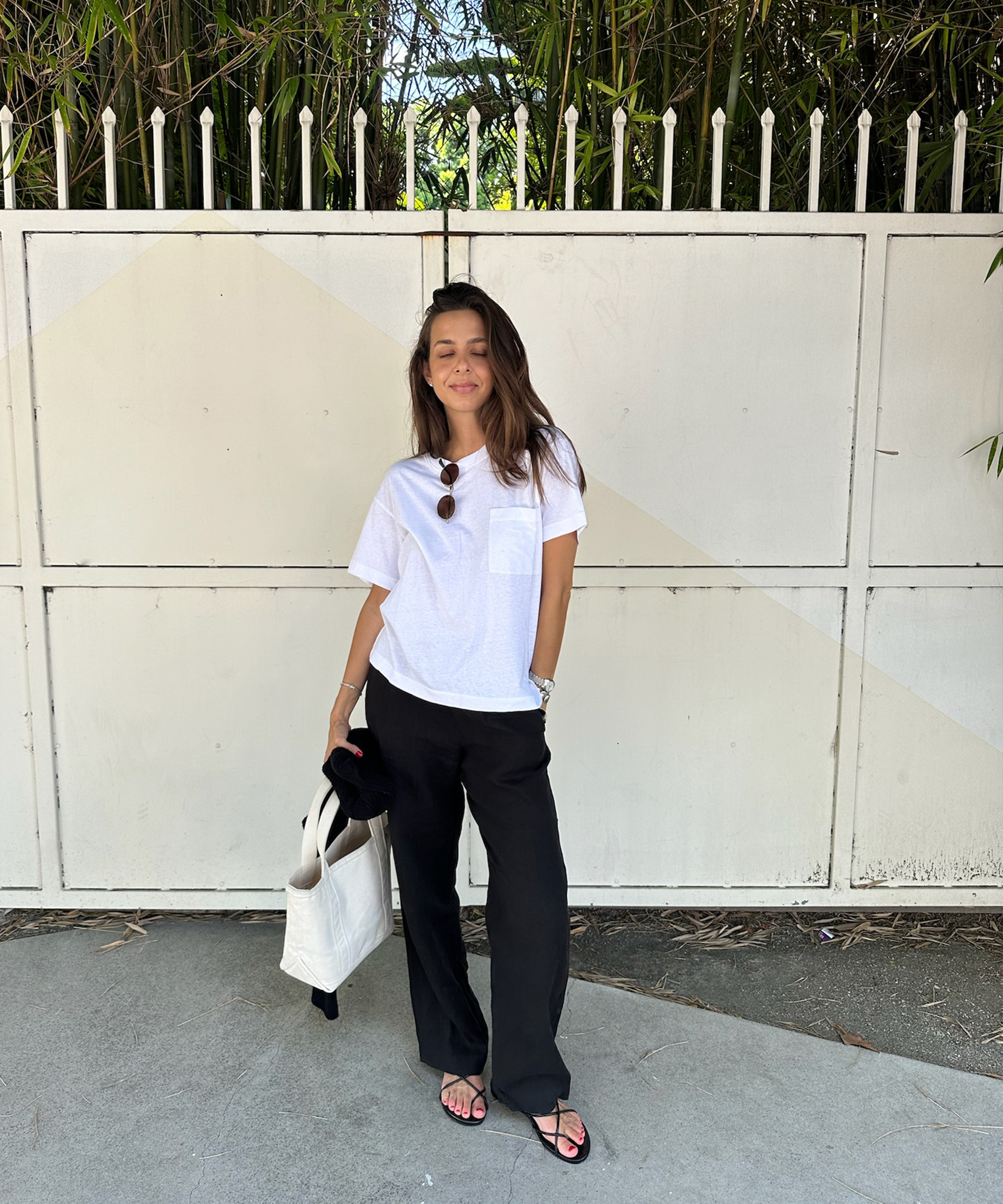 Gabriella Gofis stands in front of a white gate, wearing a white t-shirt, black pants, and sandals. She holds a white tote bag with sunglasses on her shirt, framed by tall green plants behind her.