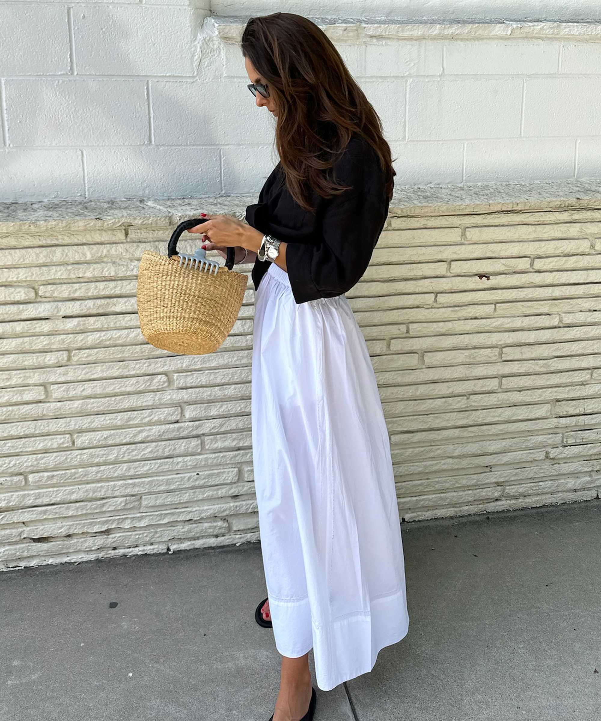 gabriella gofis standing in front of an ivory brick wall wearing a white maxi shirt and black linen button up with black sandals and a rattan bag