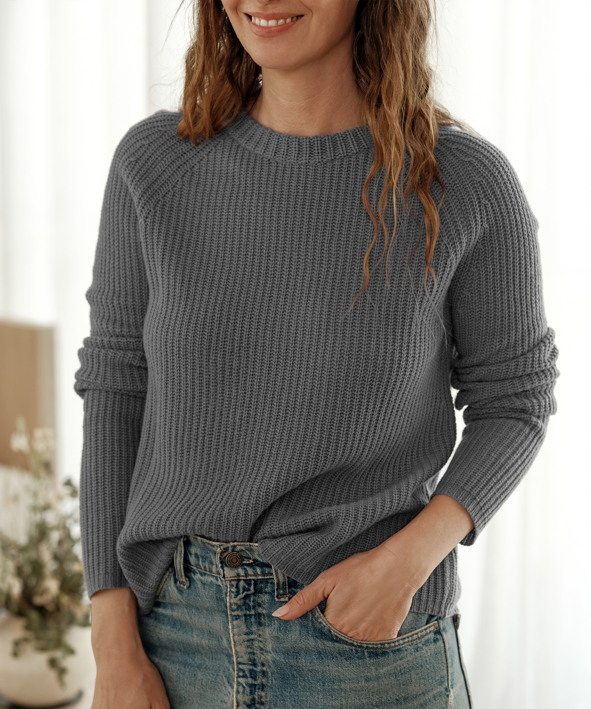 A woman with wavy hair is smiling and wearing a gray knit sweater and blue jeans, a classic example of easy outfit formulas. She stands indoors with one hand in her pocket, soft light, and blurred plants in the background.