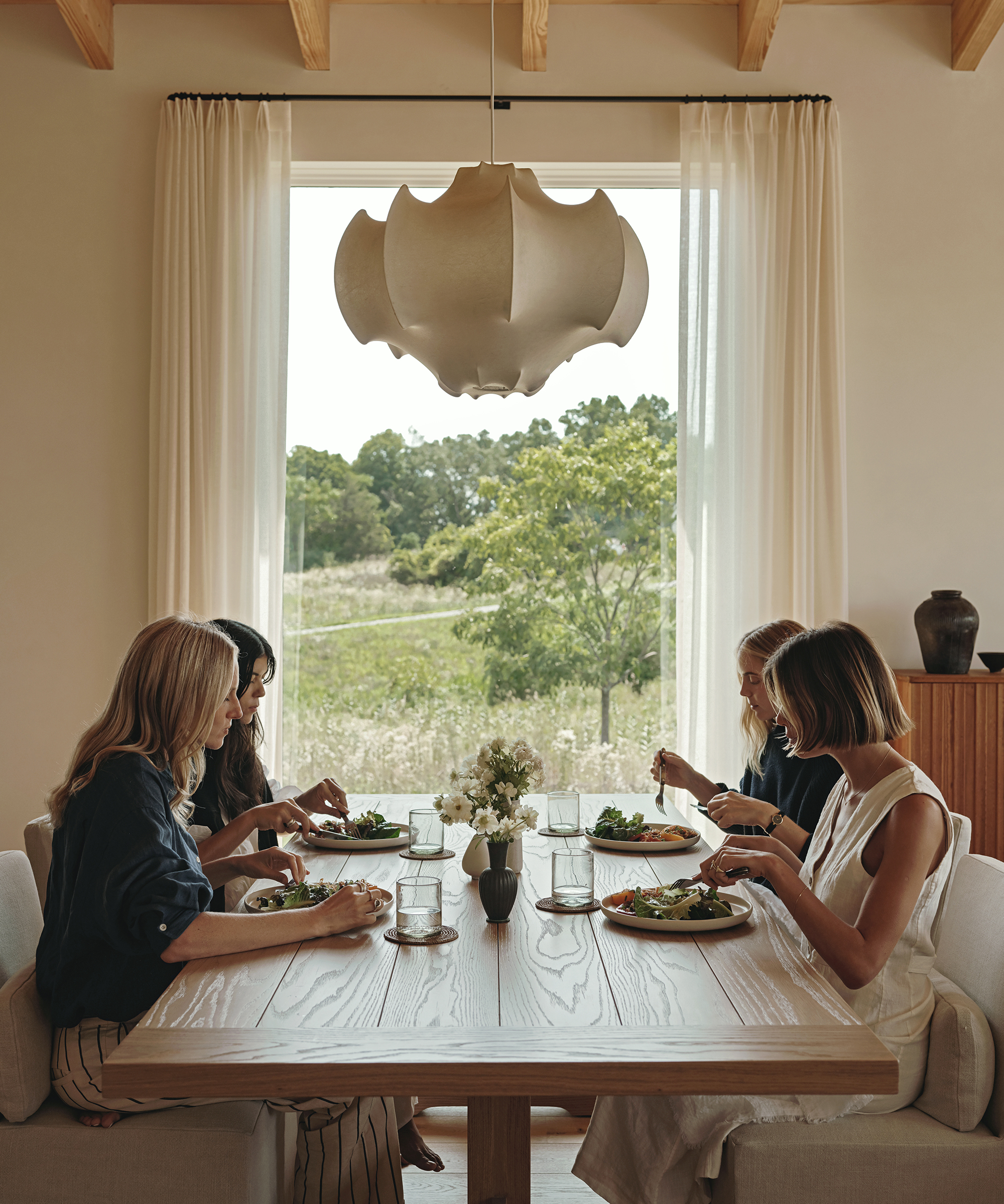 Five women sit around a wooden dining table, enjoying a meal together in a bright room with large windows, sheer curtains, and a modern pendant light—a true Jenni Kayne farmhouse vibe with greenery visible outside.