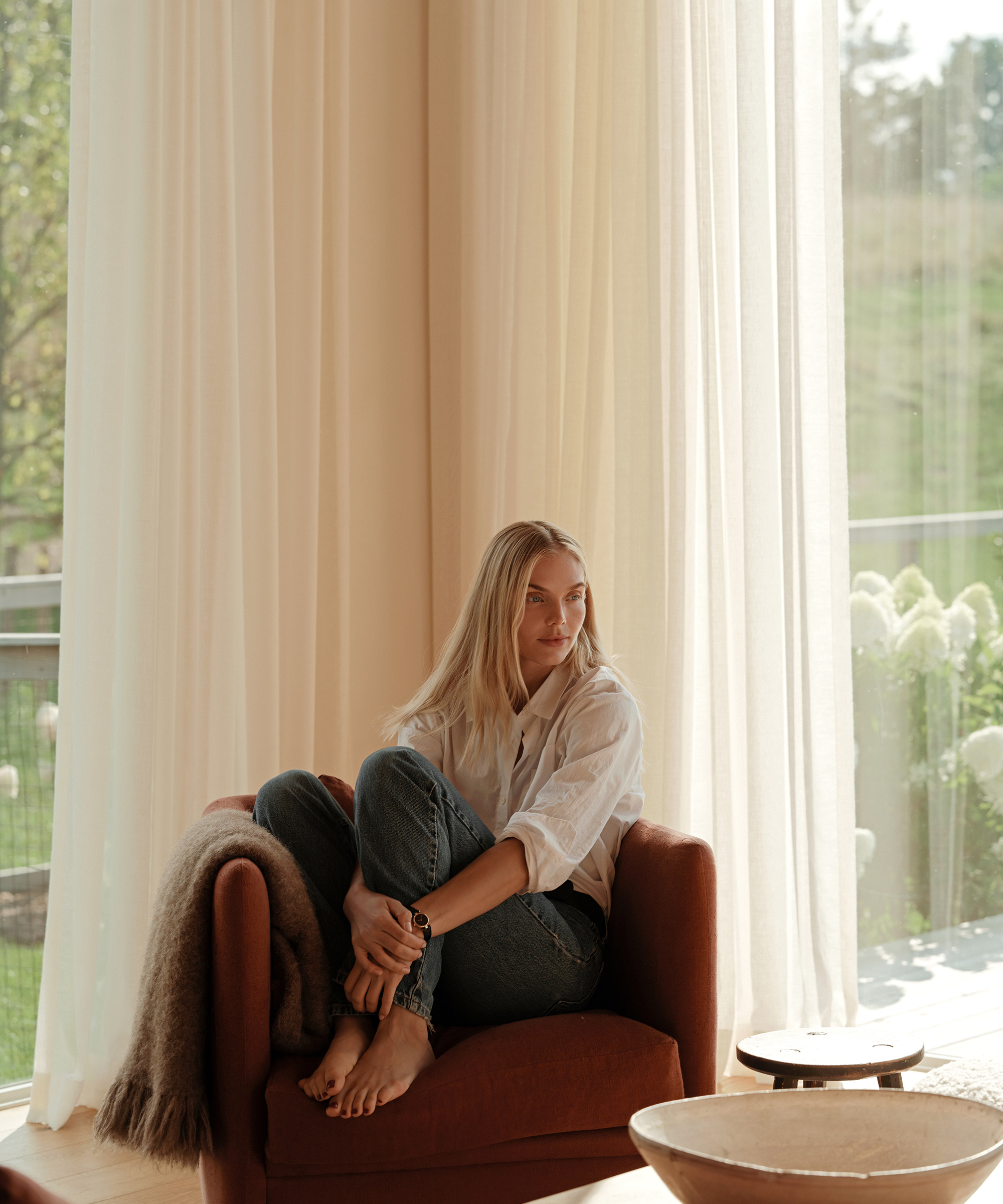 a blonde woman wearing a button down and blue jeans sitting in a red upholstered chair
