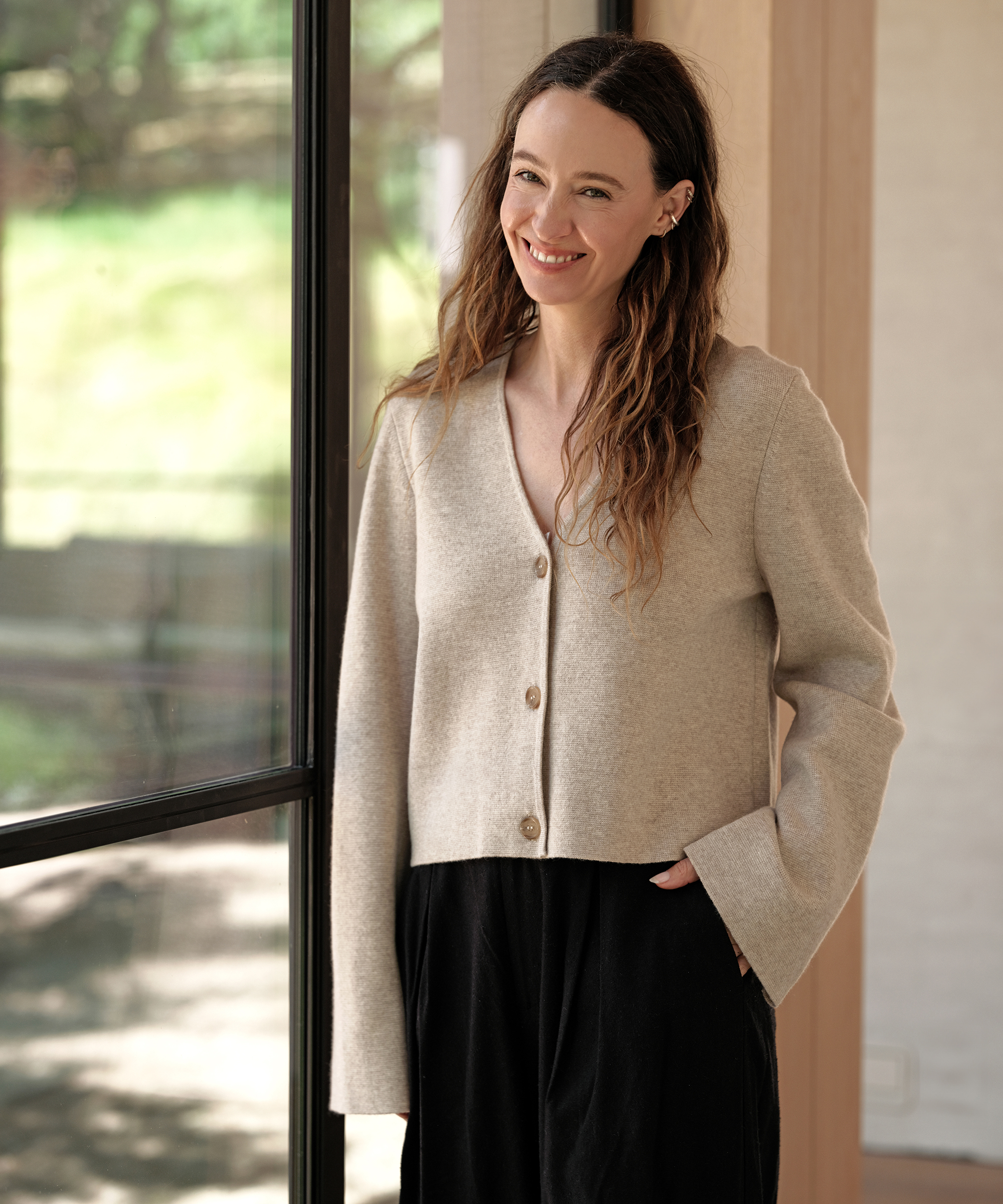 jenni leaning against a glass window wearing an oatmeal cardigan and black trousers