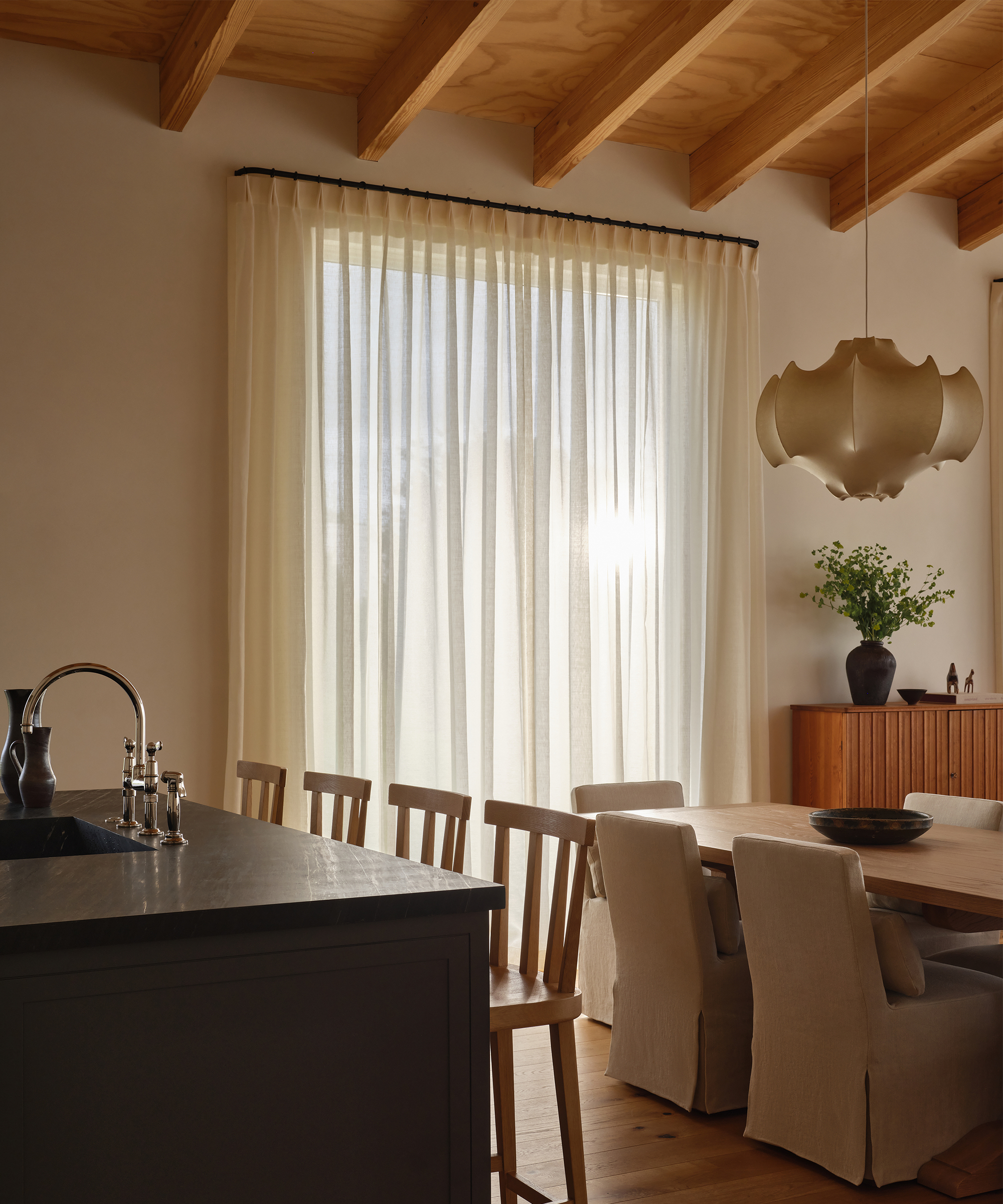 Sunlight filters through elegant window treatments in a modern dining area with a wooden table, beige upholstered chairs, a dark countertop island, and a sculptural pendant light. Wooden beams are visible on the ceiling.