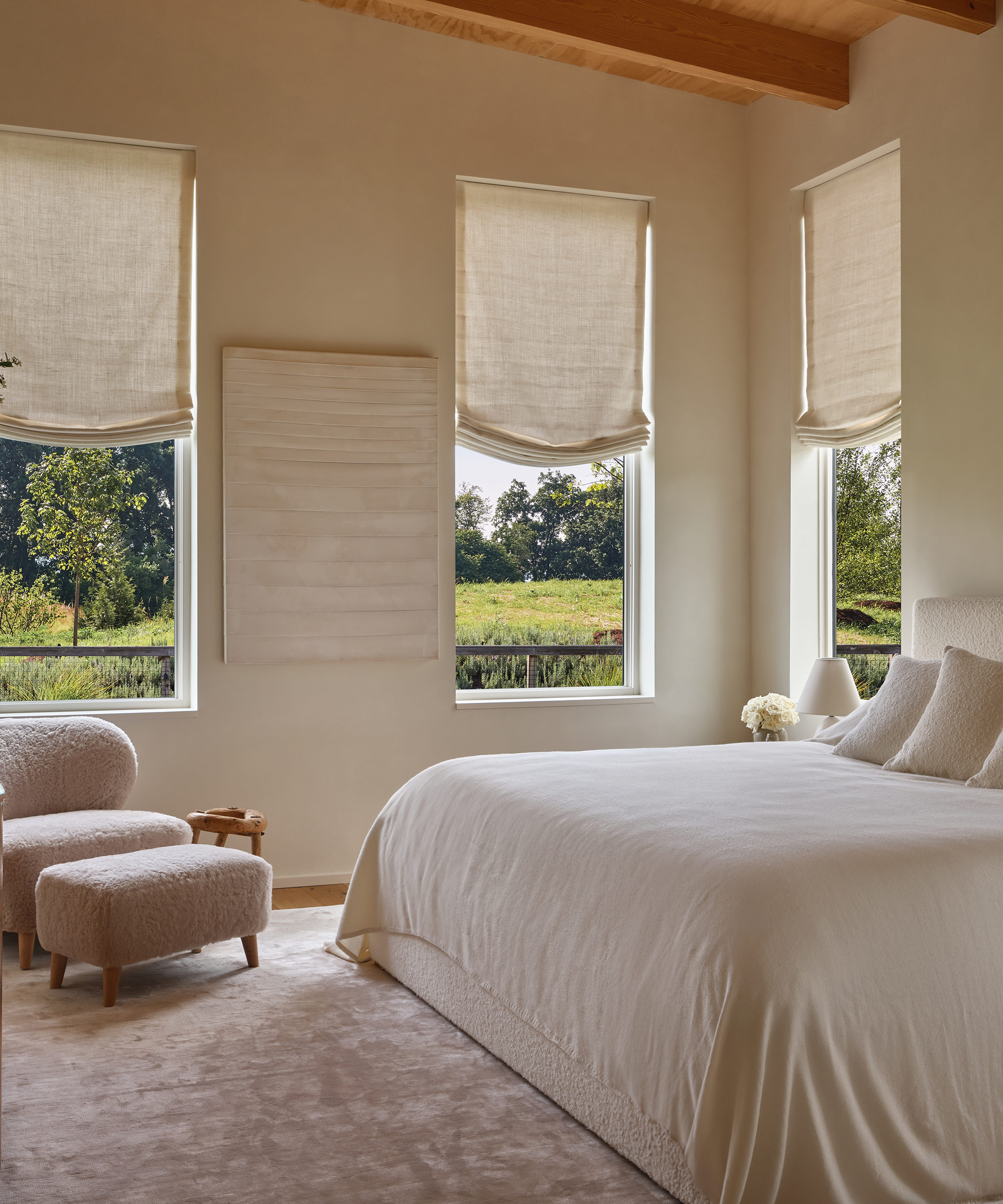 a primary bedroom with an ivory coverlet and a shearling accent chair with a shearling ottoman and plush ivory rug and ivory linen window treatments