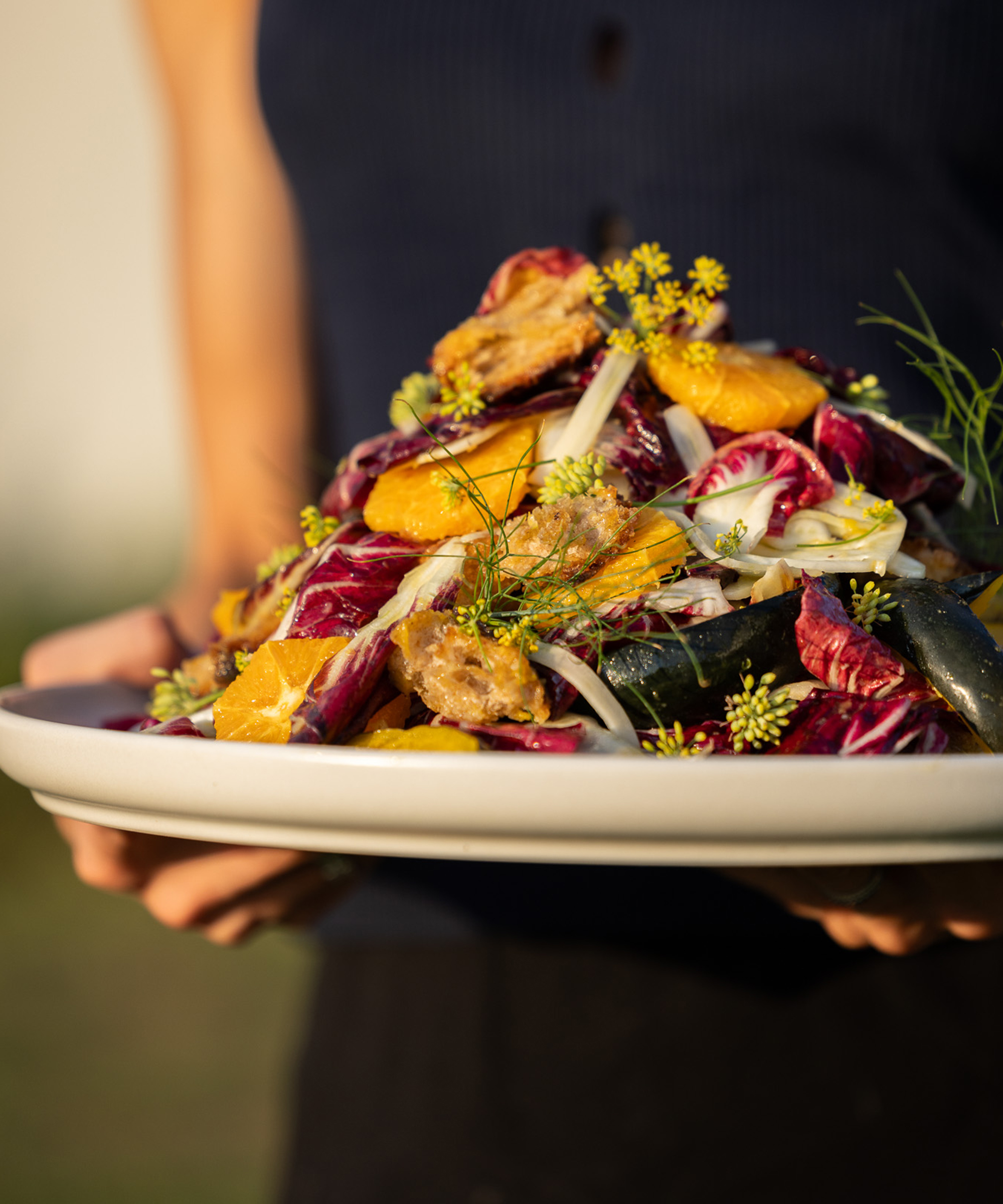A person holds a large white plate filled with a colorful salad of radicchio, orange slices, zucchini, fennel, and edible yellow flowers, with sunlight highlighting the vibrant ingredients.