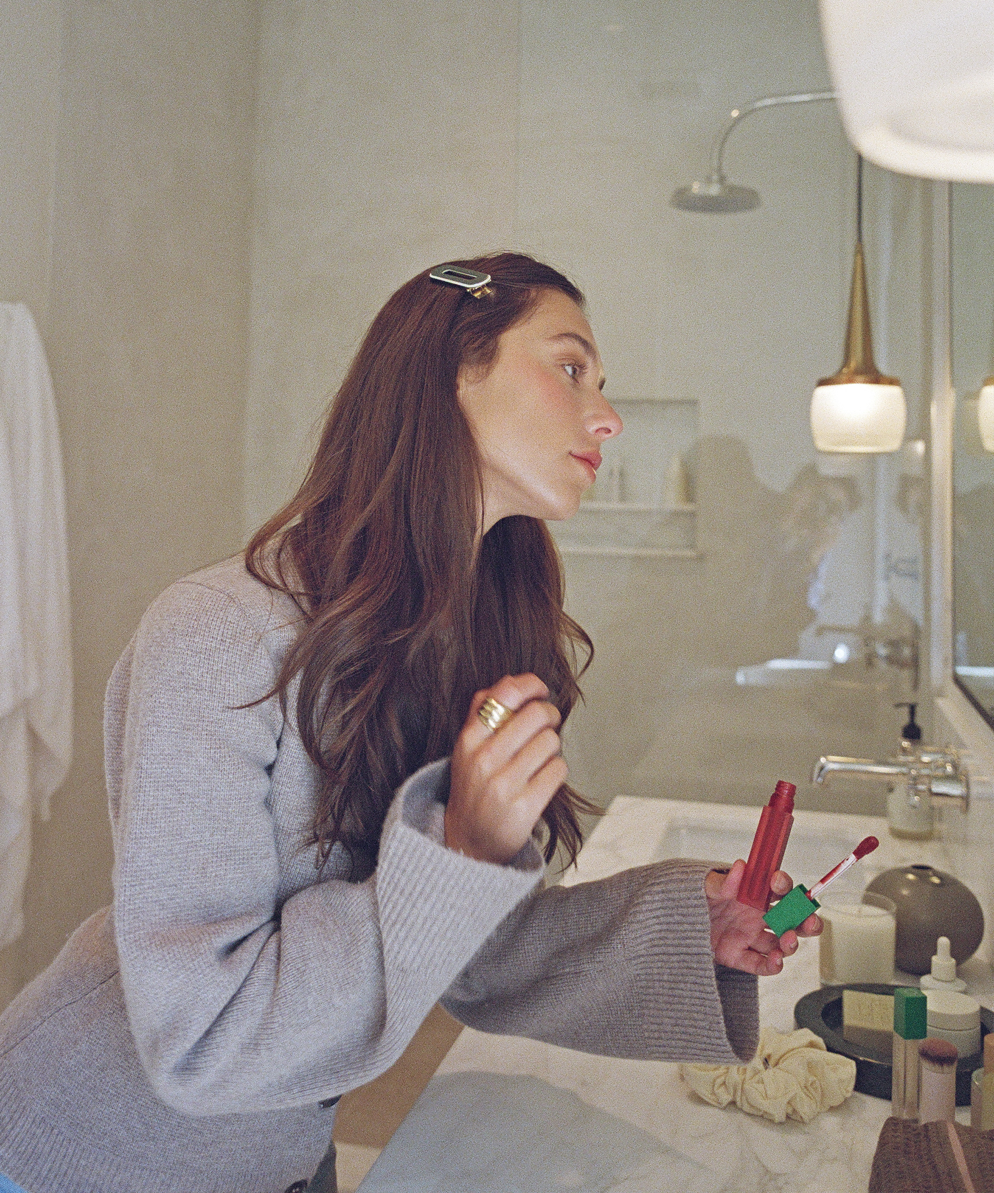 Ella McFadin, with long brown hair, applies lipstick while leaning over a bathroom counter in a gray sweater. She looks into the mirror, holding a lipstick tube, with toiletries and a towel in the background.