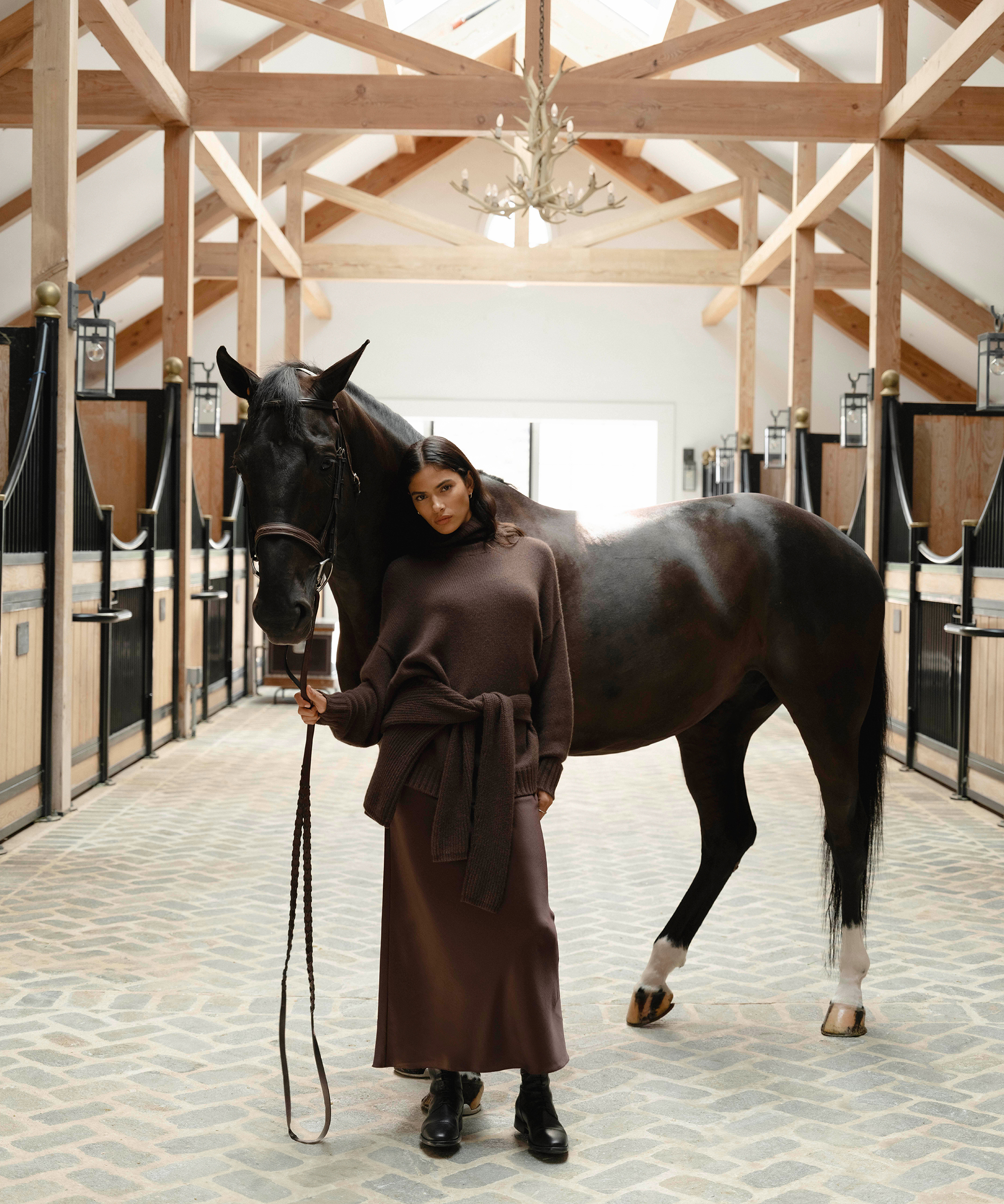 a woman wearing a brown pleated skirt and brown cashmere sweater standing next to a horse in a stable