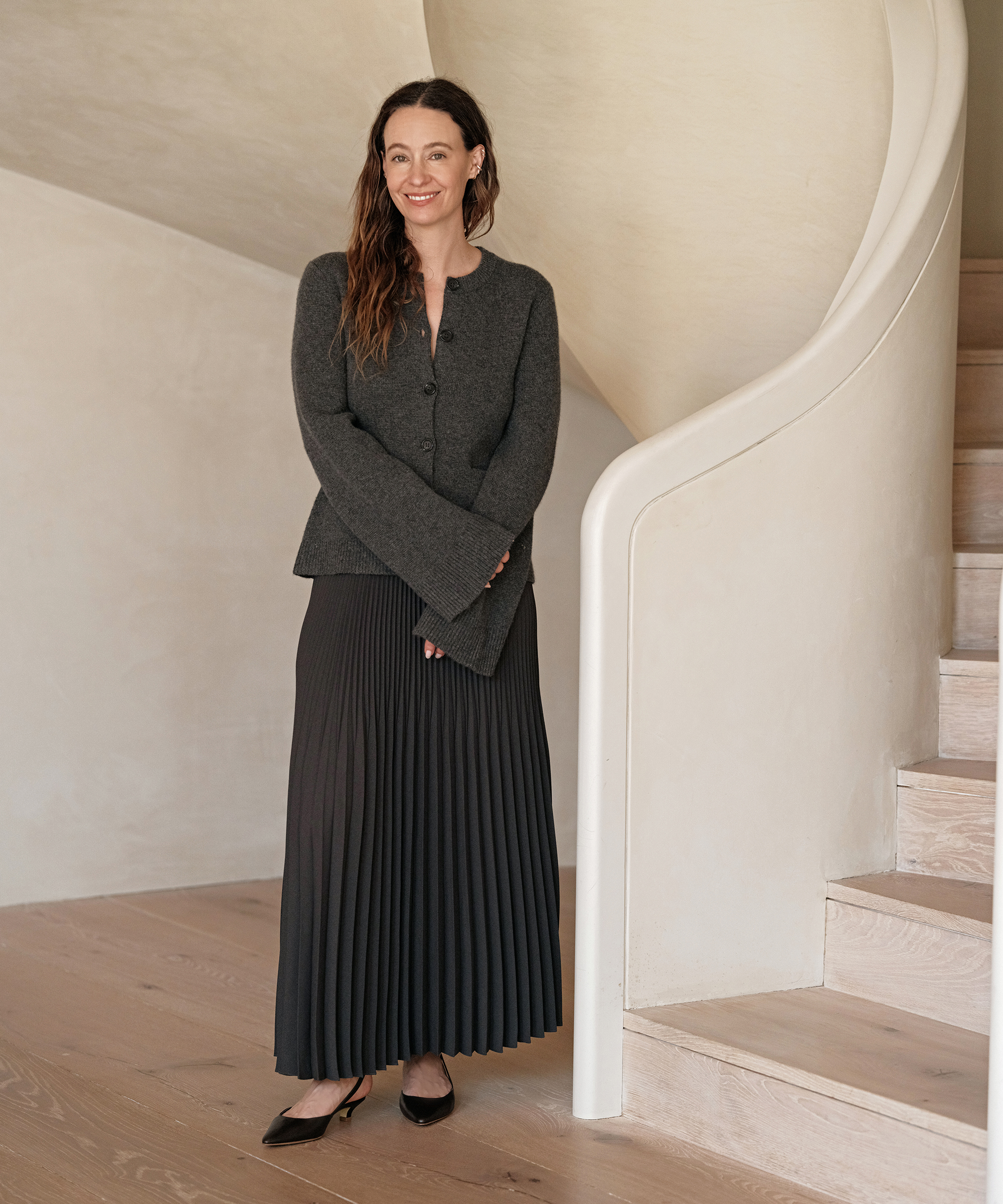 A woman with long brown hair, wearing a dark gray cardigan and a black pleated skirt, stands smiling beside a curved staircase on light wood flooring, showcasing effortless season style in a minimal, neutral-toned interior.