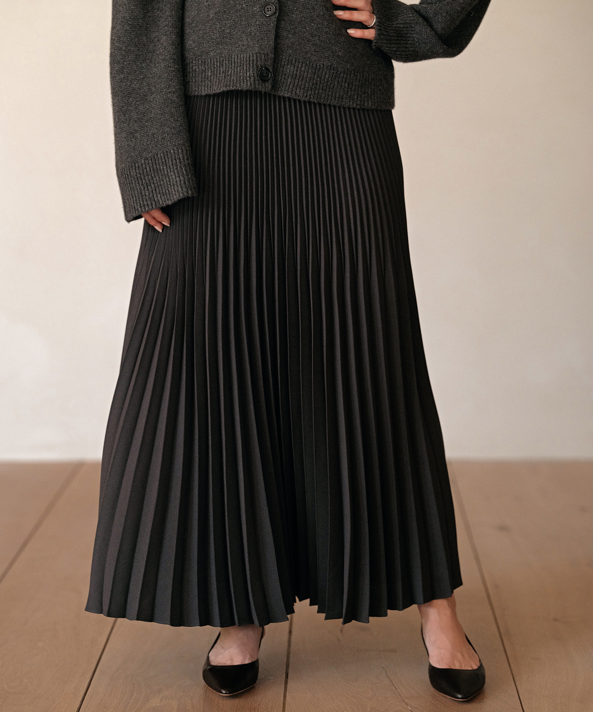 A person showcases season style in a dark pleated maxi skirt, gray knit cardigan, and black pointed-toe flats while standing on a wooden floor. The persons upper body is partially out of frame.