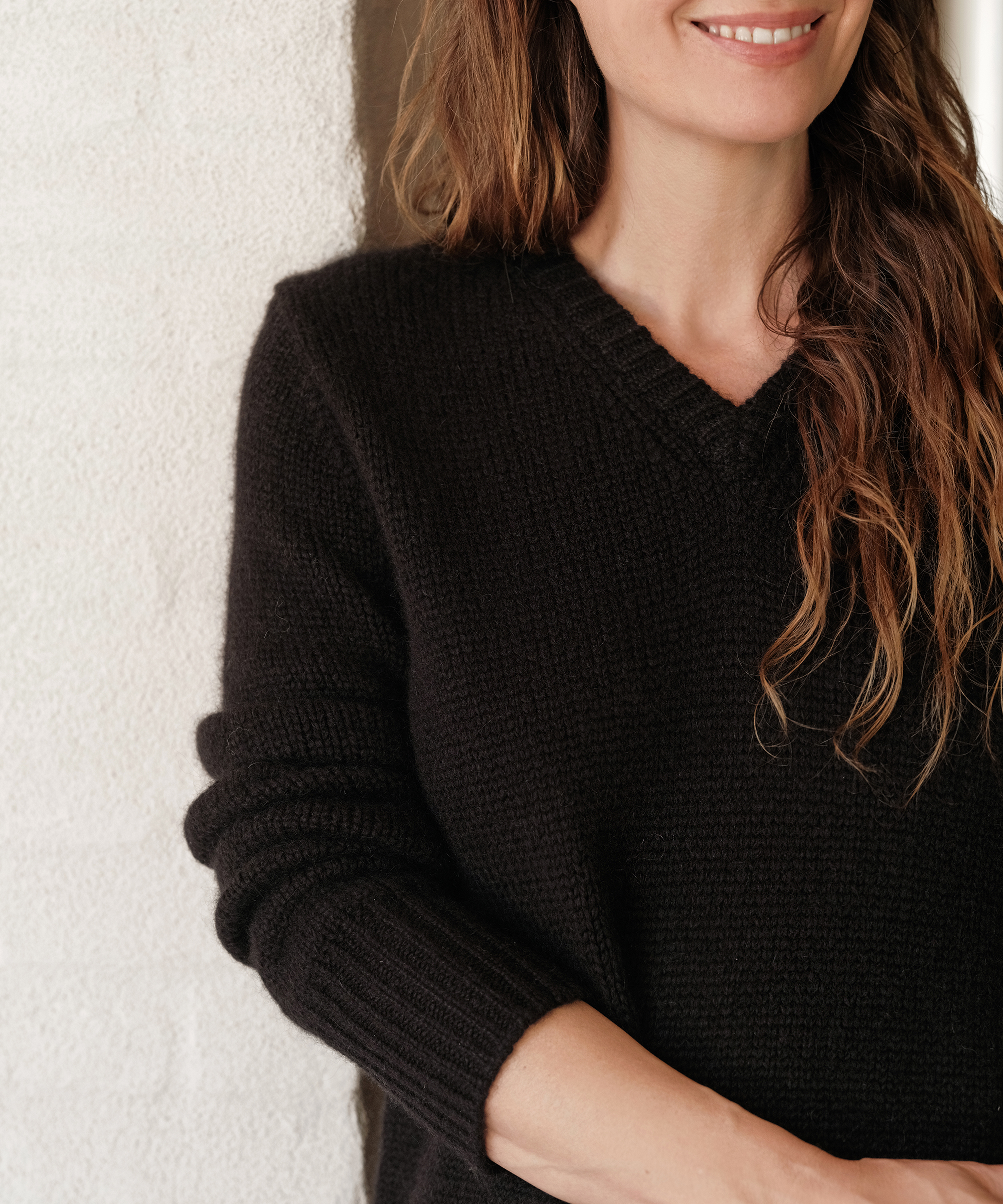 A woman with wavy brown hair, wearing a black V-neck sweater, stands against a white textured wall, smiling softly. Her chic season style is evident, with only her upper body and part of her face visible.