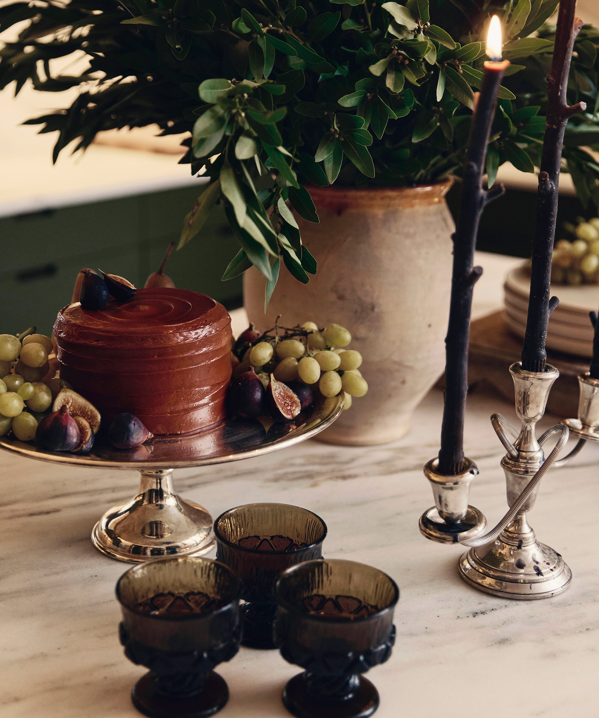 A small chocolate cake topped with figs sits on a silver stand beside bunches of grapes, dark glass goblets, two lit black candles in silver holders, and a large vase of green foliage on a marble table, evoking Augusta Hoffman’s elegant still lifes.