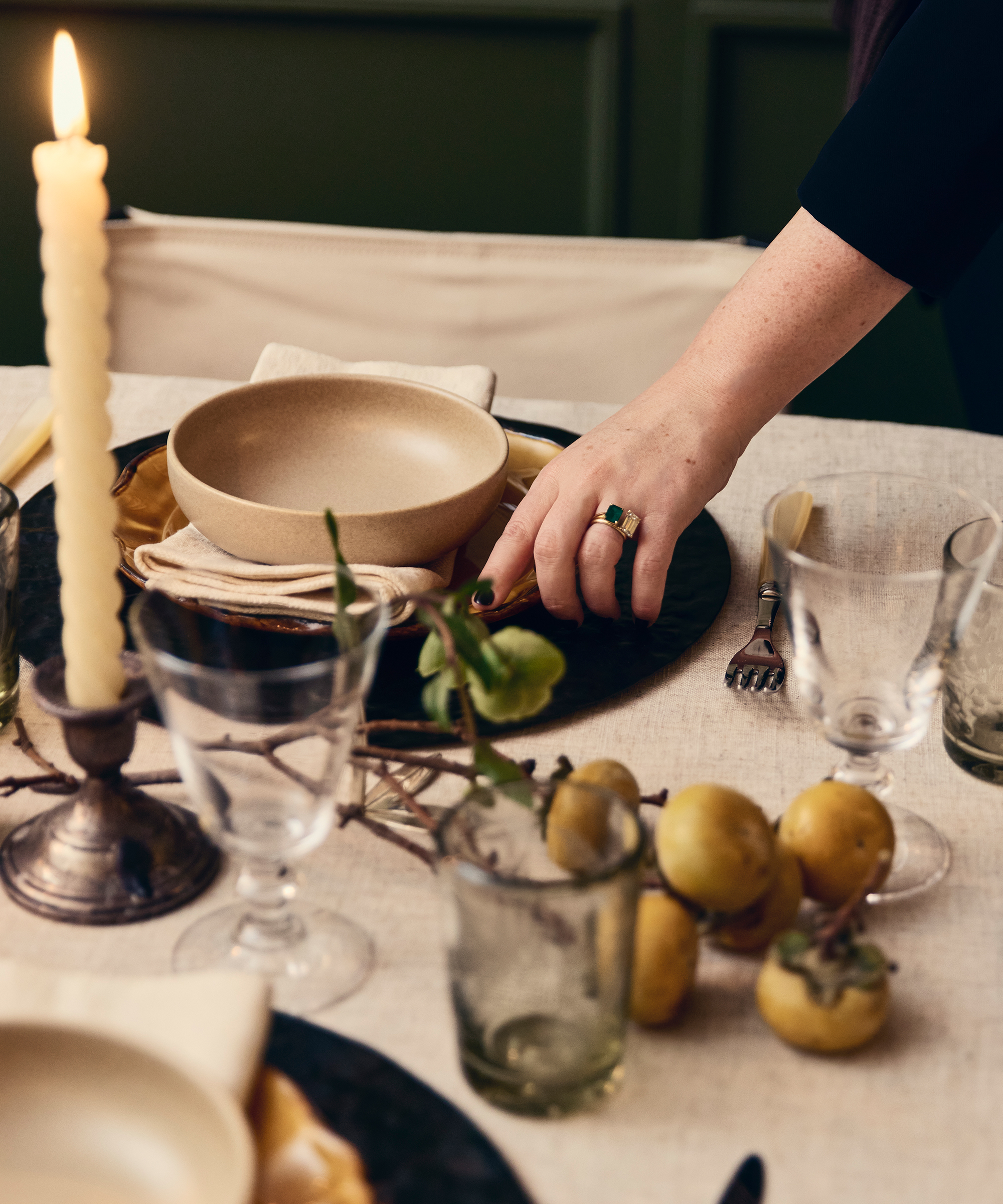 A hand with a green ring arranges a bowl on a set dining table styled by Augusta Hoffman, featuring a lit candle, glassware, napkins, cutlery, and a bunch of persimmons on a cream-colored tablecloth.