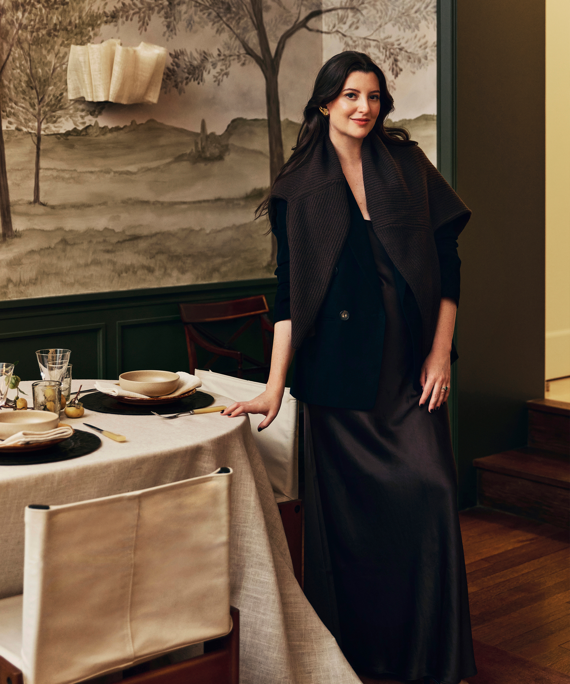 augusta hoffman wearing a brown silk dress and brown sweater over the shoulders standing next to a dining table with a linen tablecloth