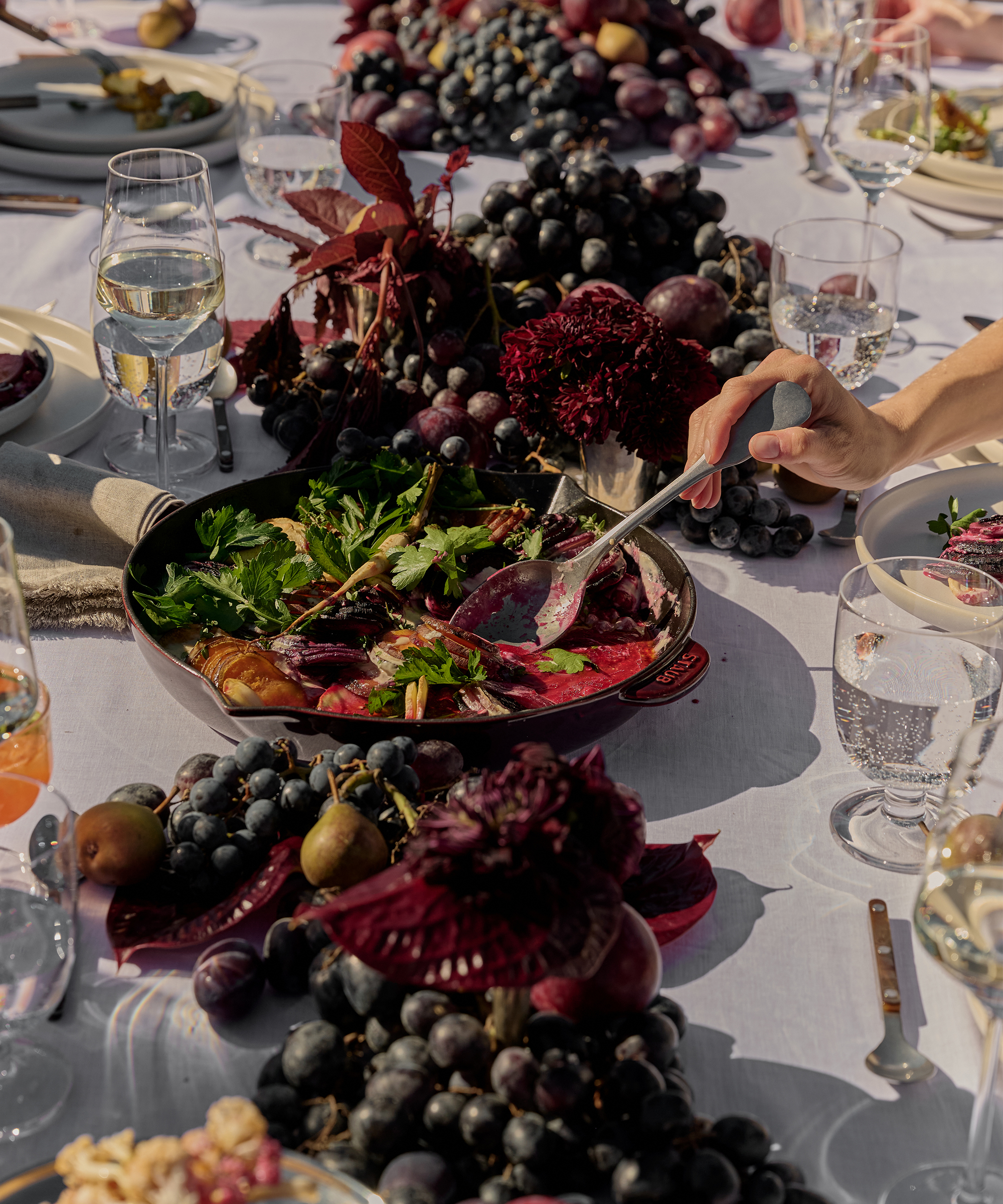 A hand serves salad from a Staub pan on a festively set table decorated with grapes, dark flowers, and glassware, creating an elegant outdoor dining scene.