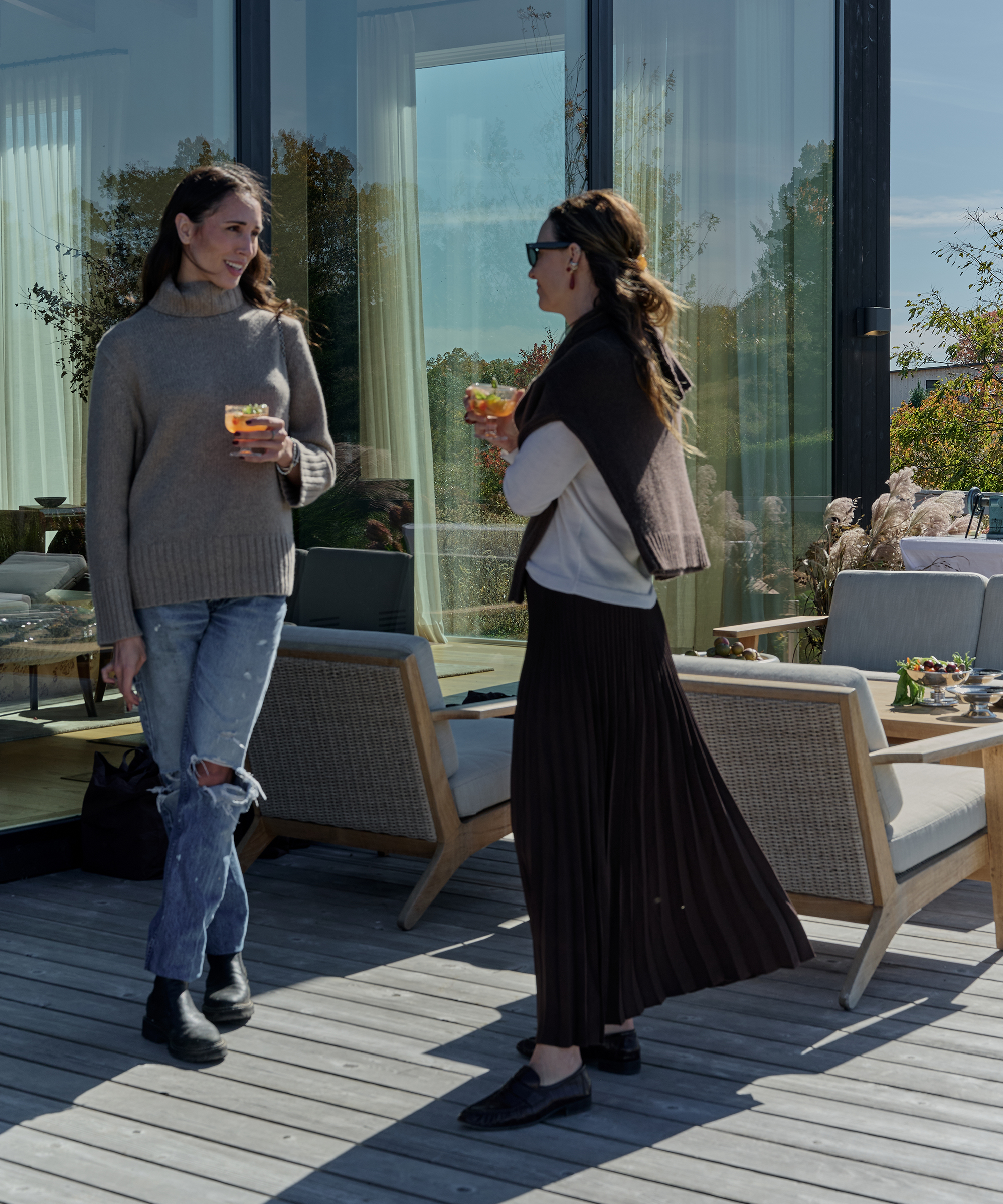 Two women stand on a sunlit outdoor patio, holding drinks and chatting beside modern outdoor furniture. A Staub cast iron pot sits on the table near them, adding a touch of style to their casual gathering.