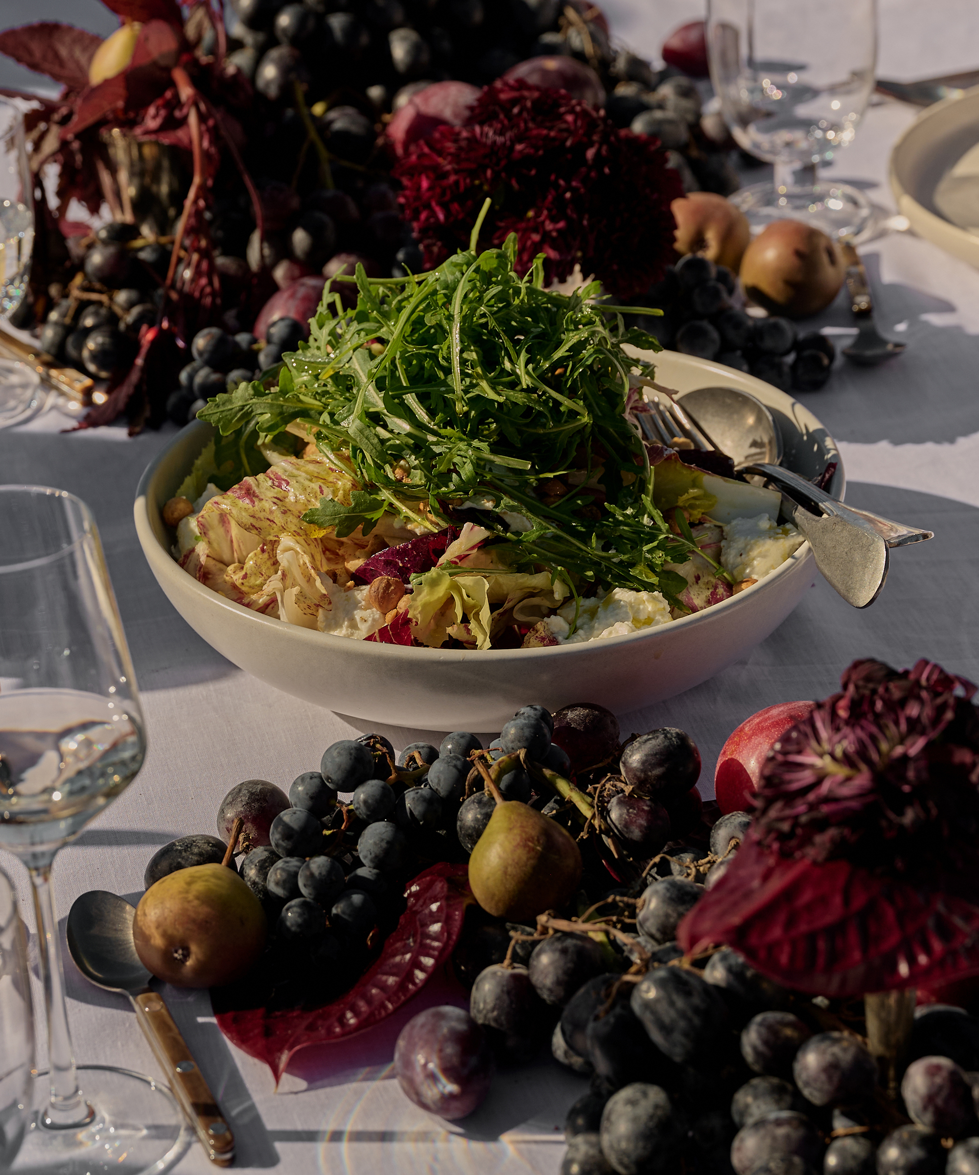 a taupe serving bowl with salad on a table with a bountiful fruit arrangement