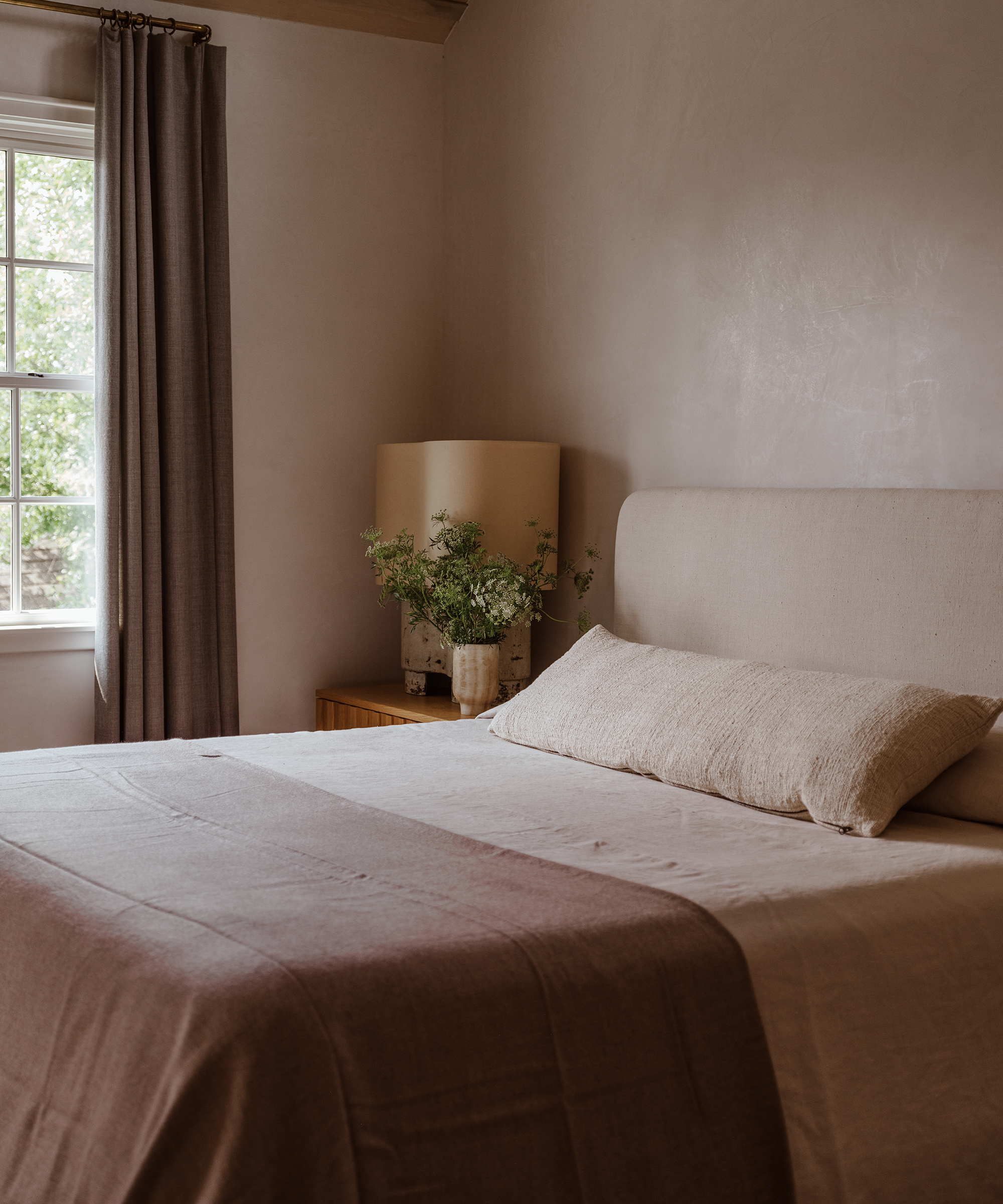 A cozy bedroom design with neutral tones features a bed with beige linens, a large textured pillow, a wooden nightstand holding a lamp and vase of green foliage, and a window with gray curtains letting in natural light.