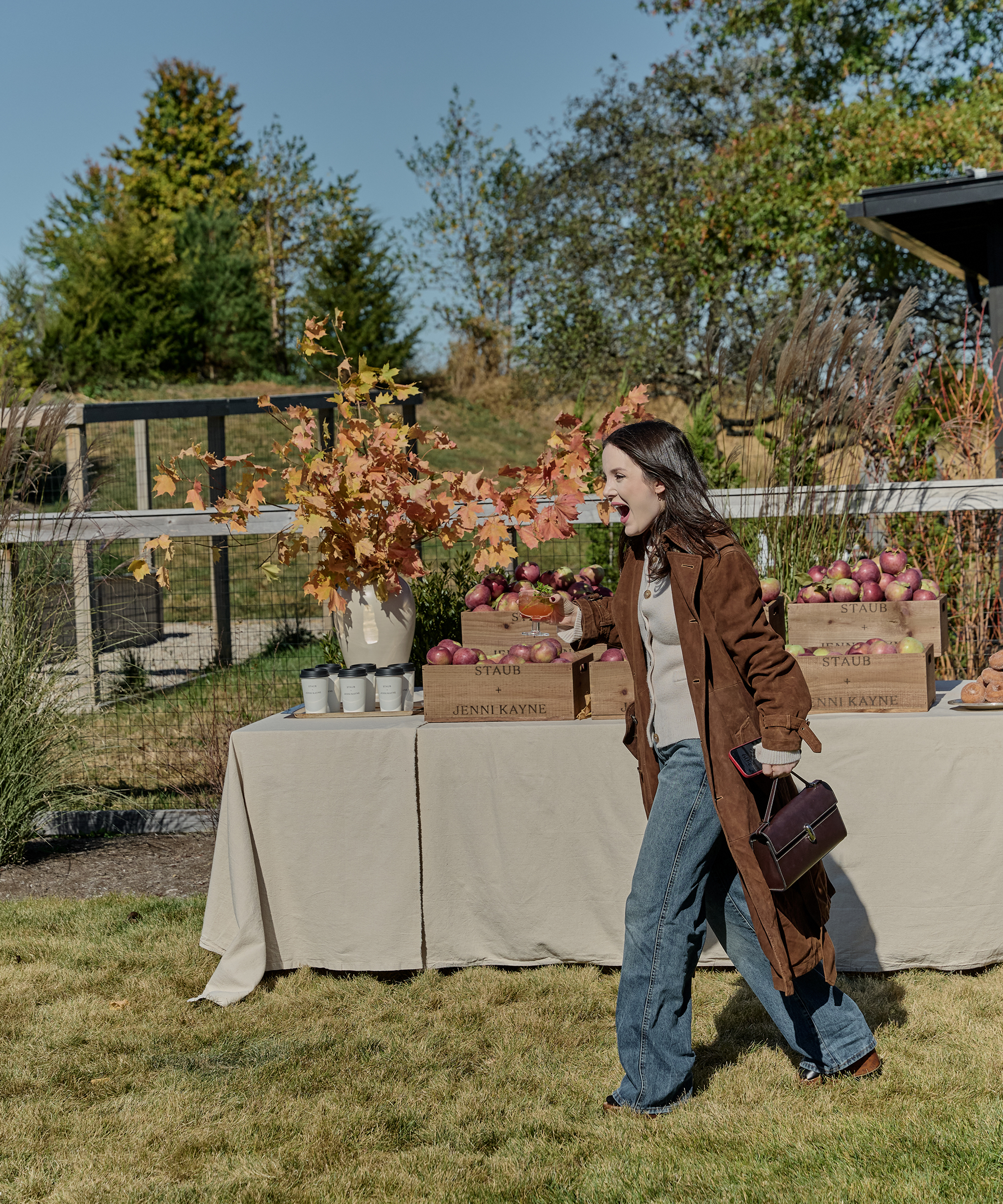 a woman running in the grass wearing jeans and a trench coat in front of a table with a linen tablecloth