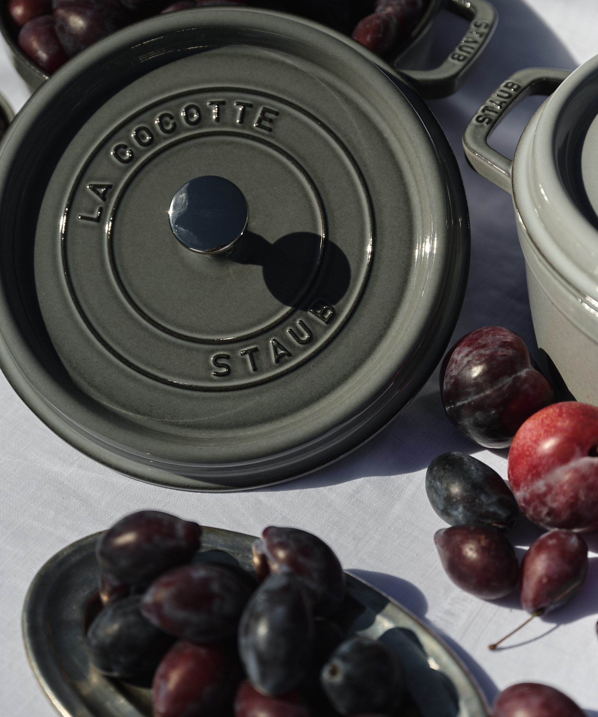 A grey Staub cocotte with a lid reading La Cocotte Staub sits on a white surface beside another pot and a metal plate filled with fresh plums and grapes, showcasing the timeless elegance of Staub cookware.