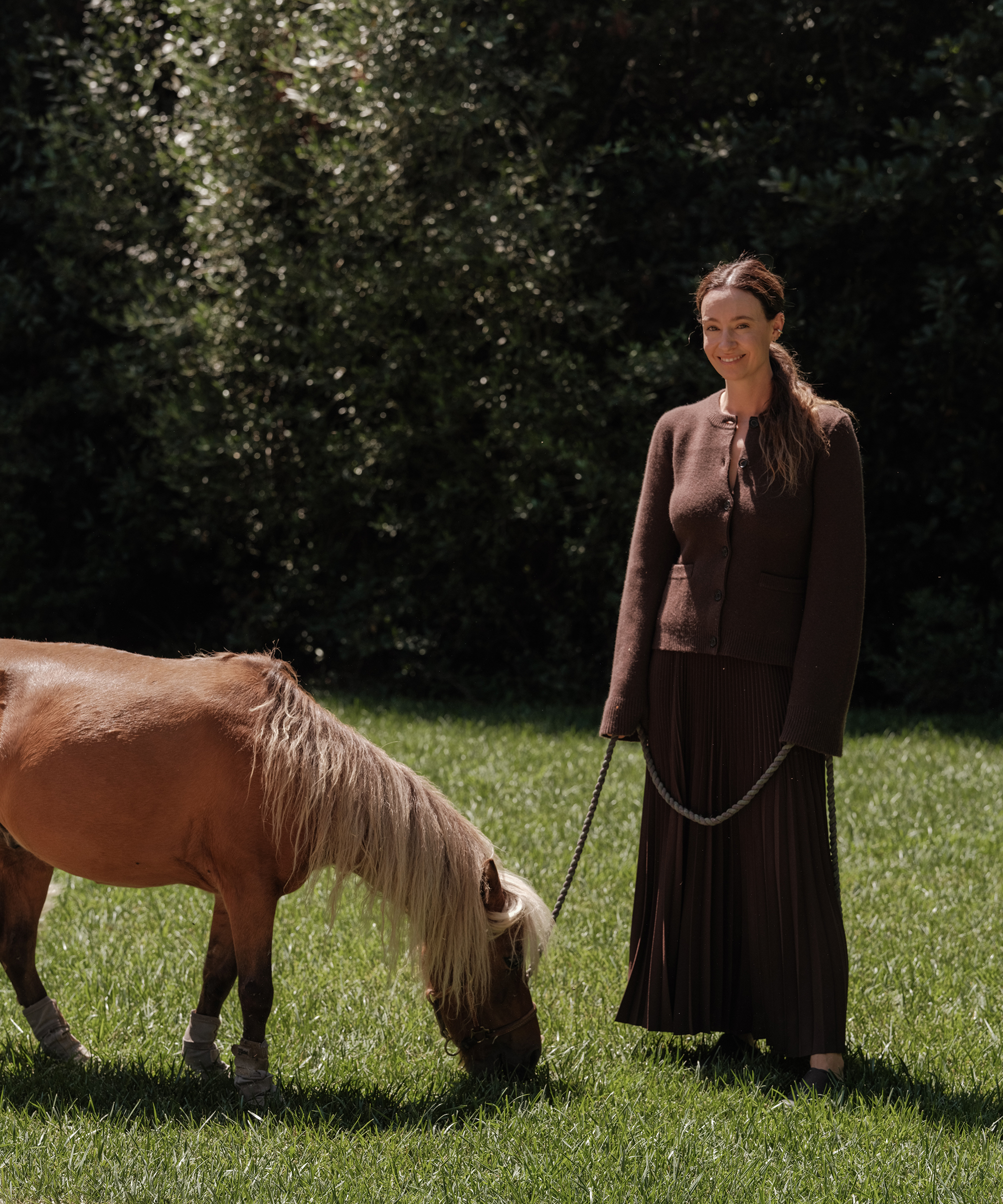 jenni walking in her backyard with a horse wearing a brown pleated skirt and brown cardigan