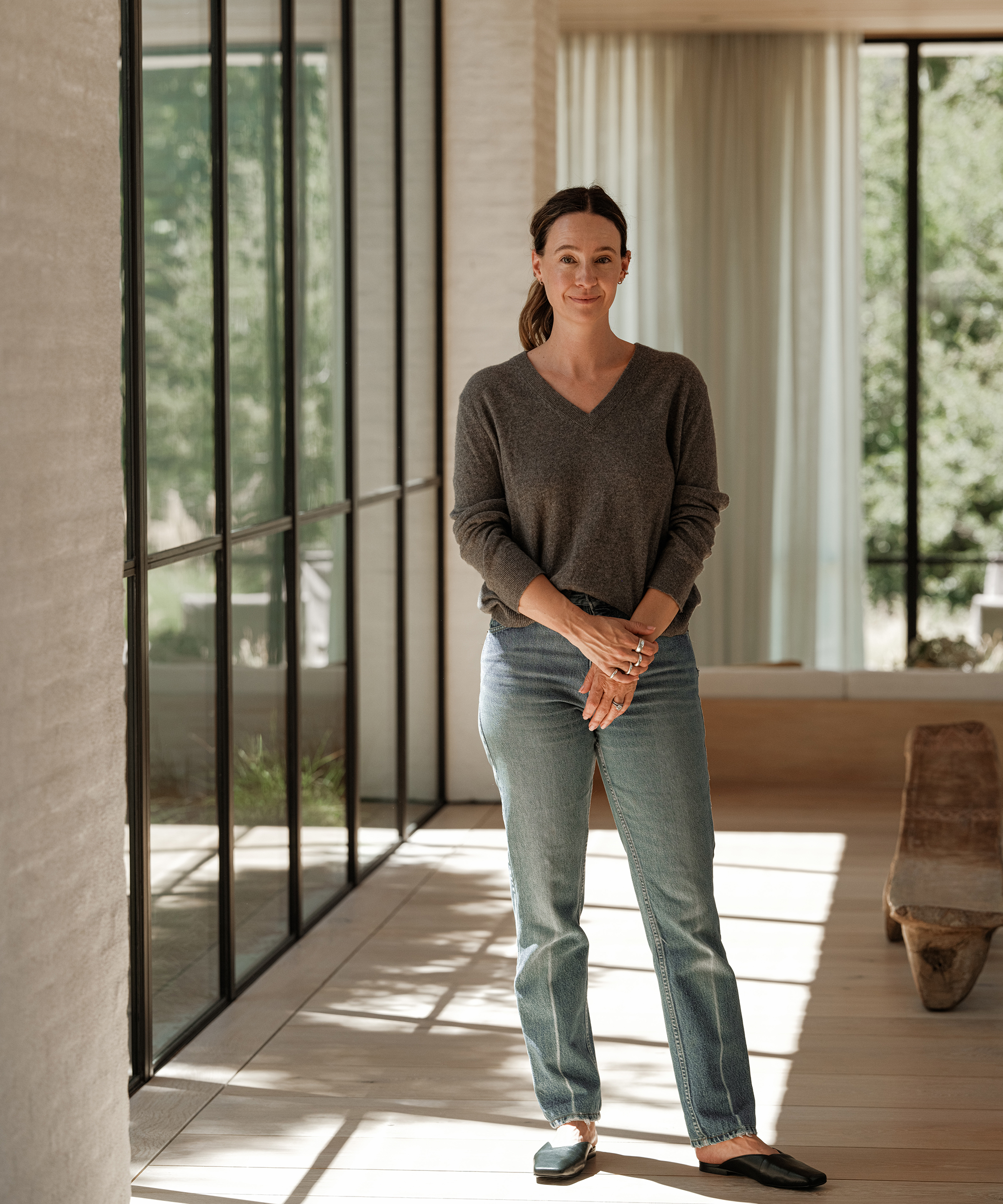 A woman stands indoors near tall windows, smiling gently in a bright, modern room with natural light. She wears a cozy fall outfit: a grey sweater, light blue jeans, and black flats, with her hands clasped in front of her.