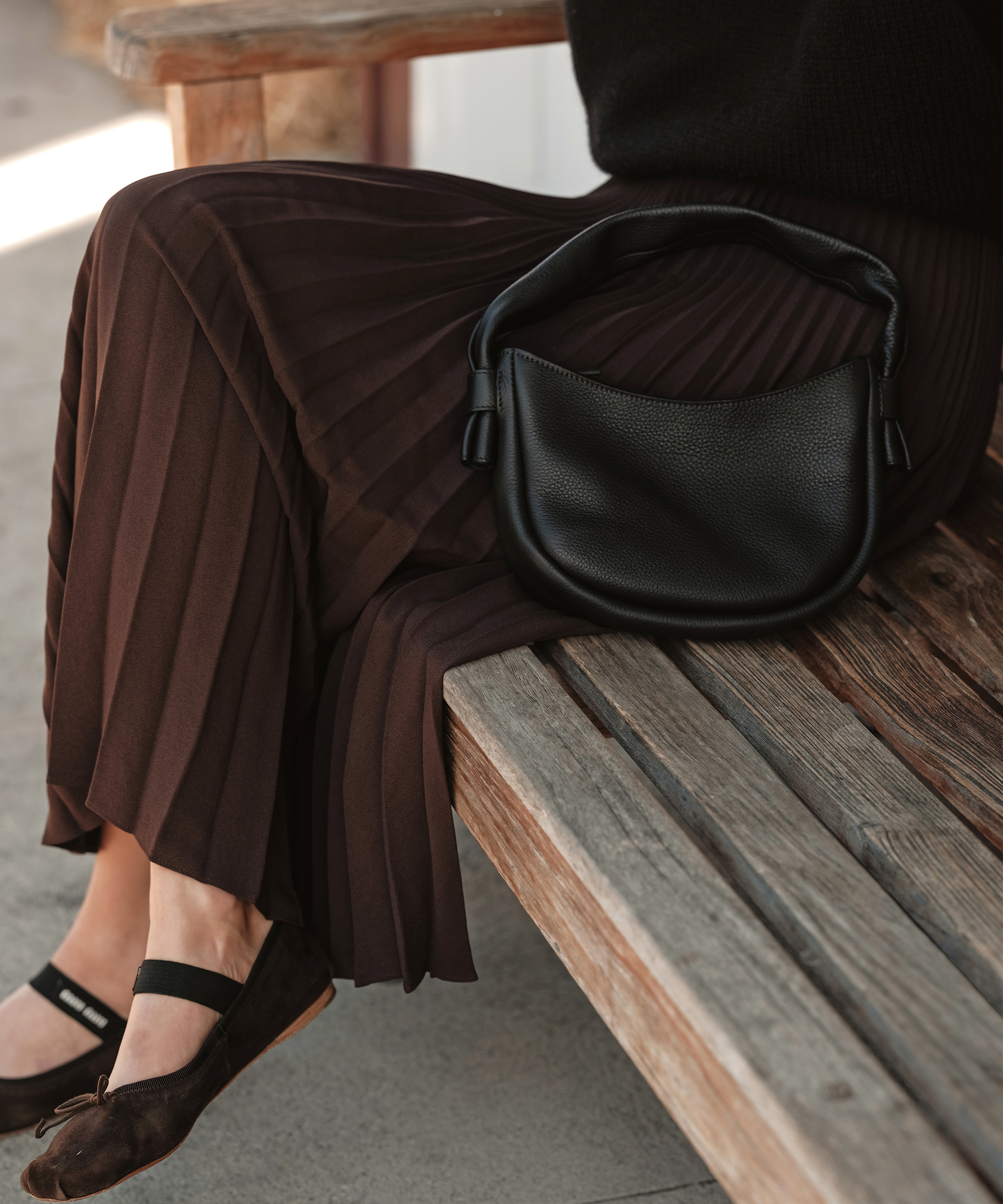a person sitting in a chocolate pleated skirt on a wooden bench with ballet flats and a black leather purse