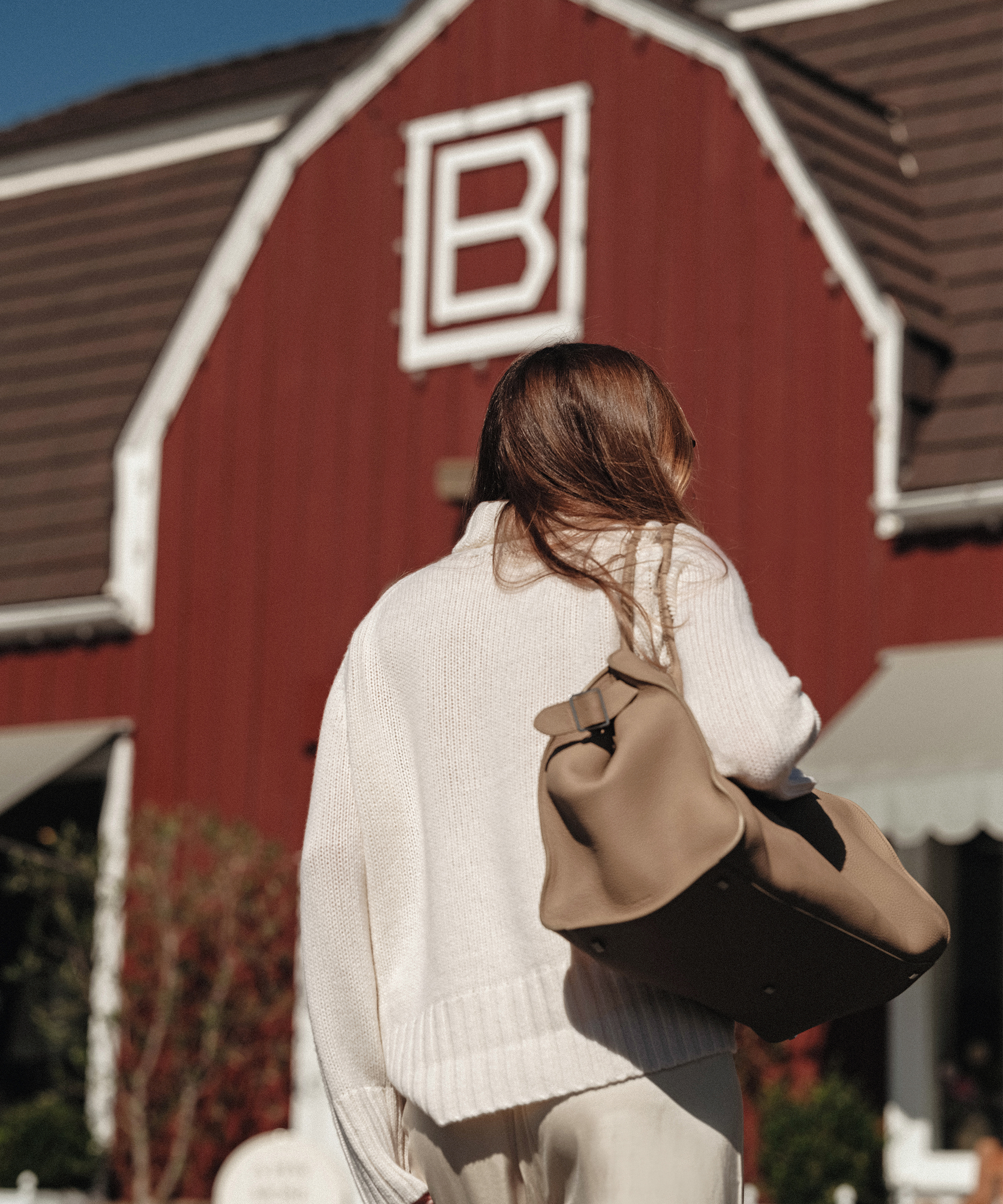 Sara Walker, with long brown hair, wearing a cream sweater and carrying a tan bag, stands in front of a red barn building with a white letter B on it.