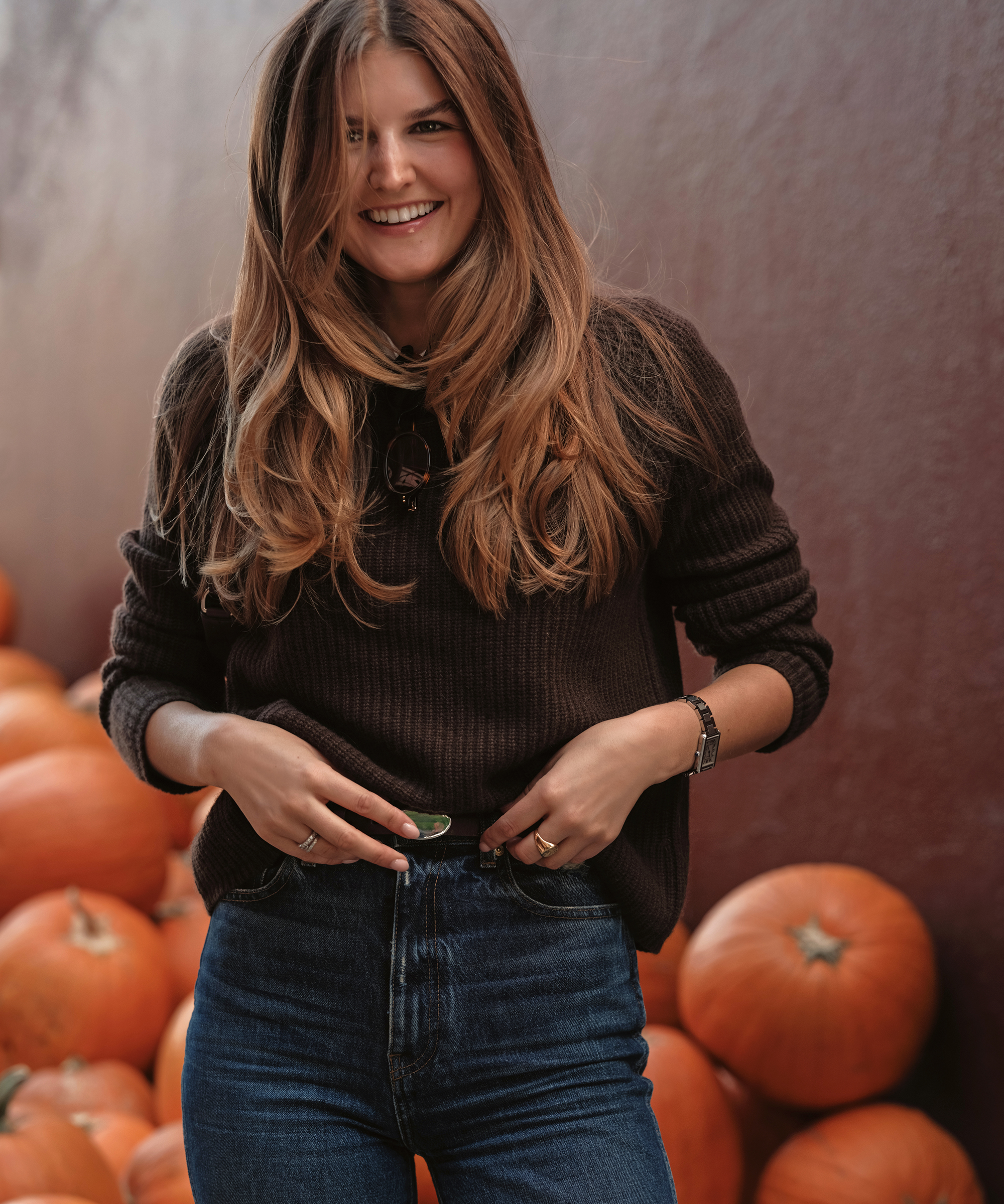 Sara Walker, with long brown hair, wearing a dark sweater and blue jeans, stands smiling in front of a pile of pumpkins against a maroon wall.