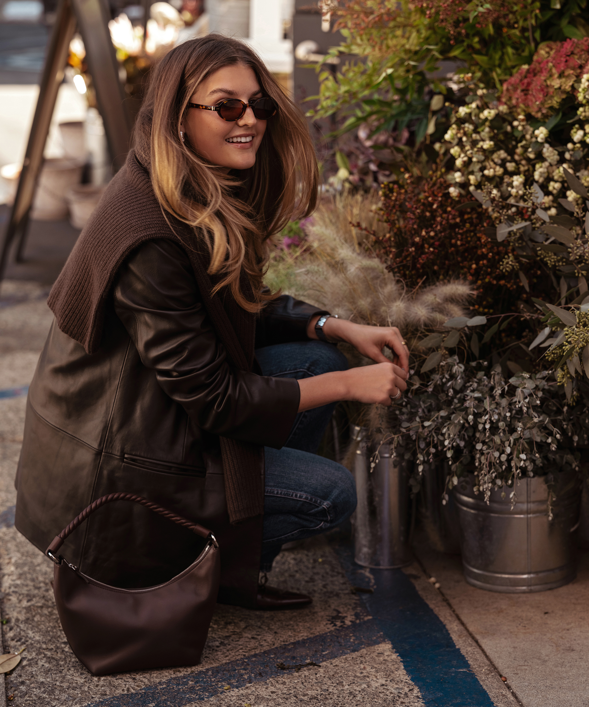 a woman wearing a chocolate leather blazer and jeans looking at buckets of flowers outside of a shop
