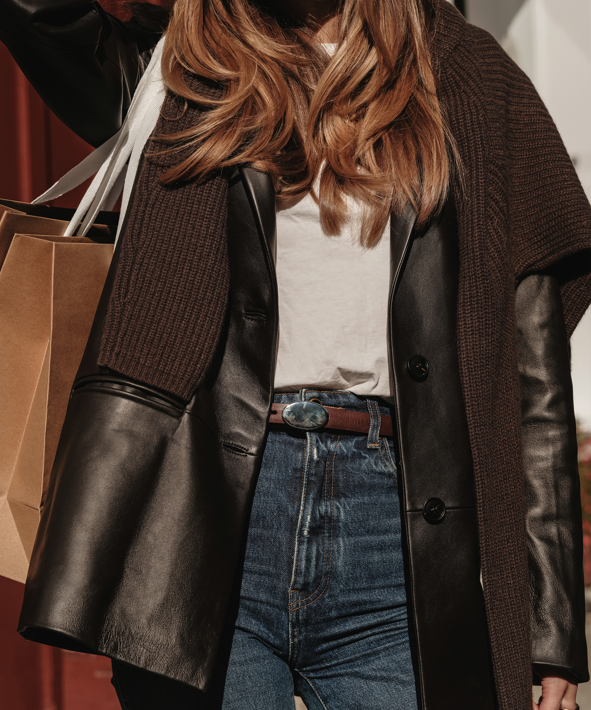 Sara Walker wears a black leather coat, white t-shirt, and blue jeans while carrying a brown shopping bag. A brown sweater is draped over her shoulders, and her face is not visible.