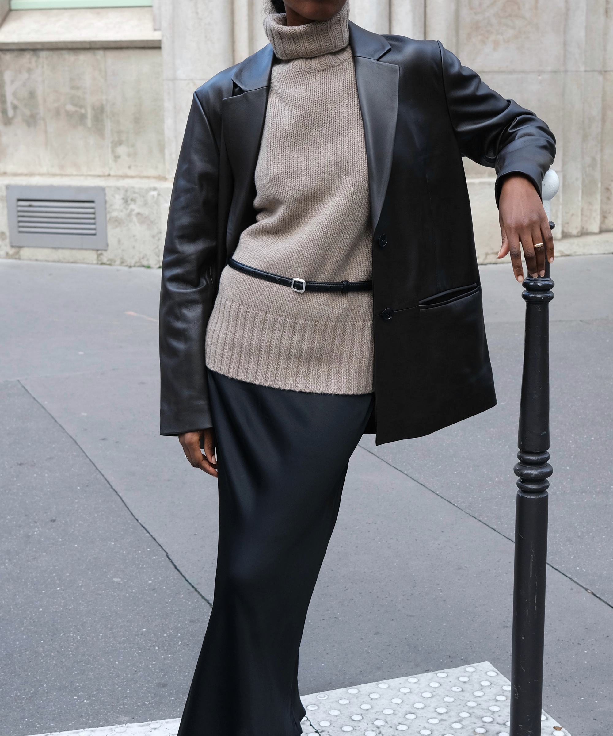 a woman in an almond cashmere turtleneck and a black silk skirt and a leather blazer leaning against a pole outside