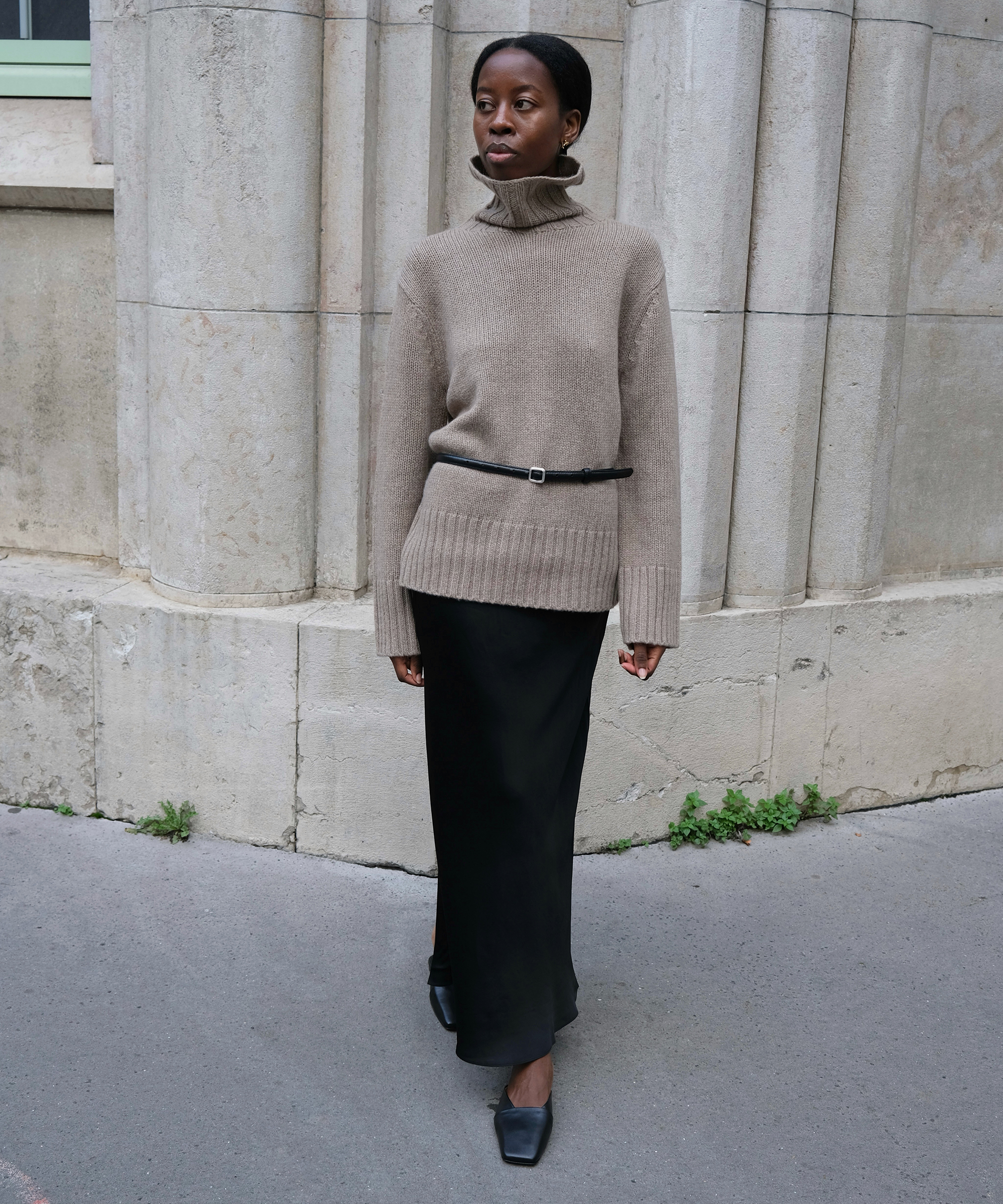 A woman stands outdoors by a stone wall, wearing a beige turtleneck sweater belted at the waist, a long black skirt, and black shoes. Channeling sylvie mus, she looks to the side with a calm expression.