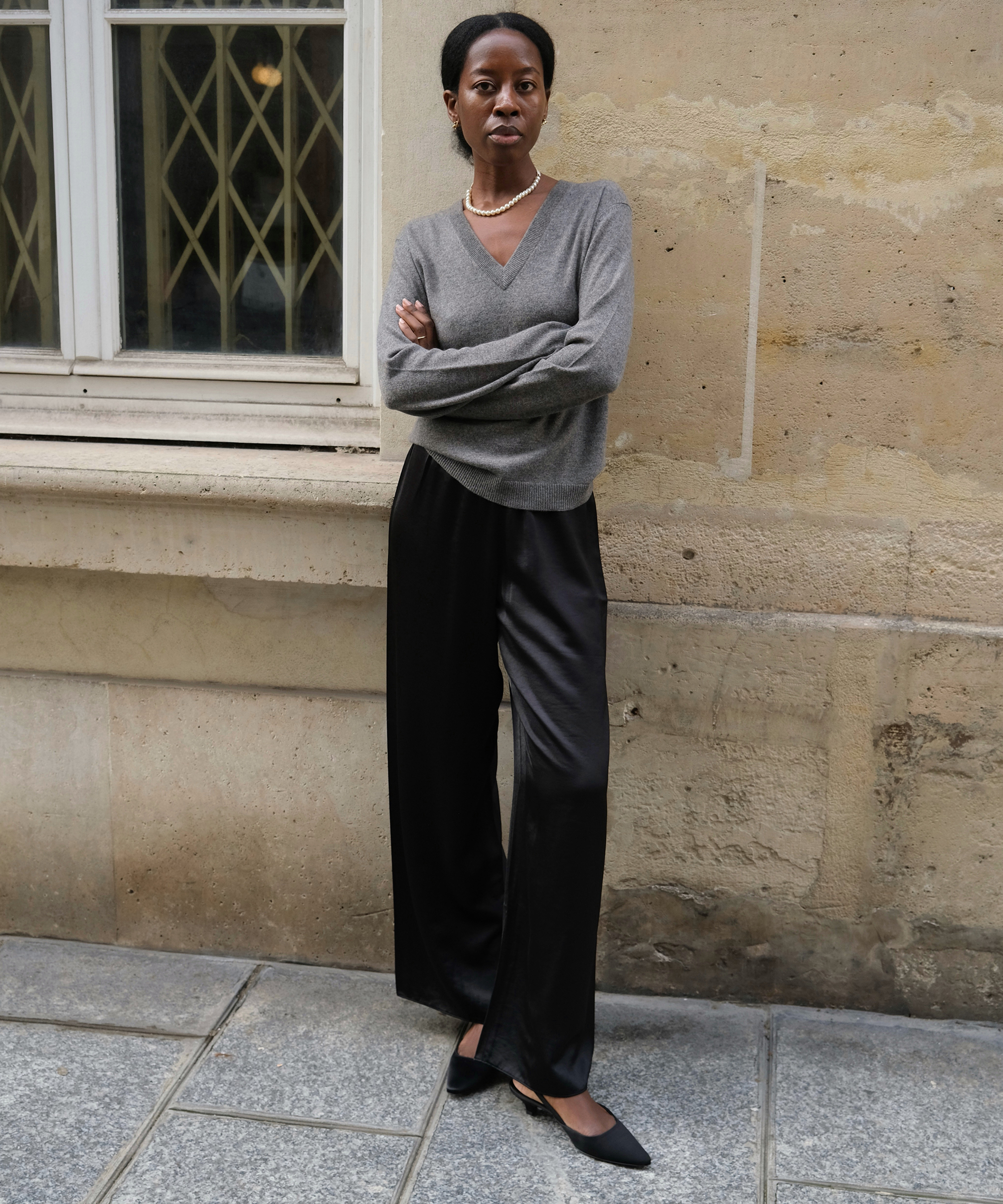 a woman standing in a gray vneck cashmere sweater and black silk pants with kitten heels standing outside