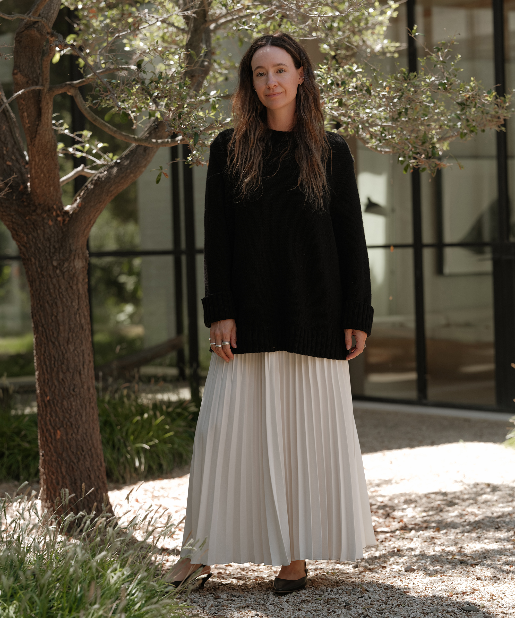 jenni wearing an ivory pleated skirt and black cashmere sweater standing outside in a gravel path