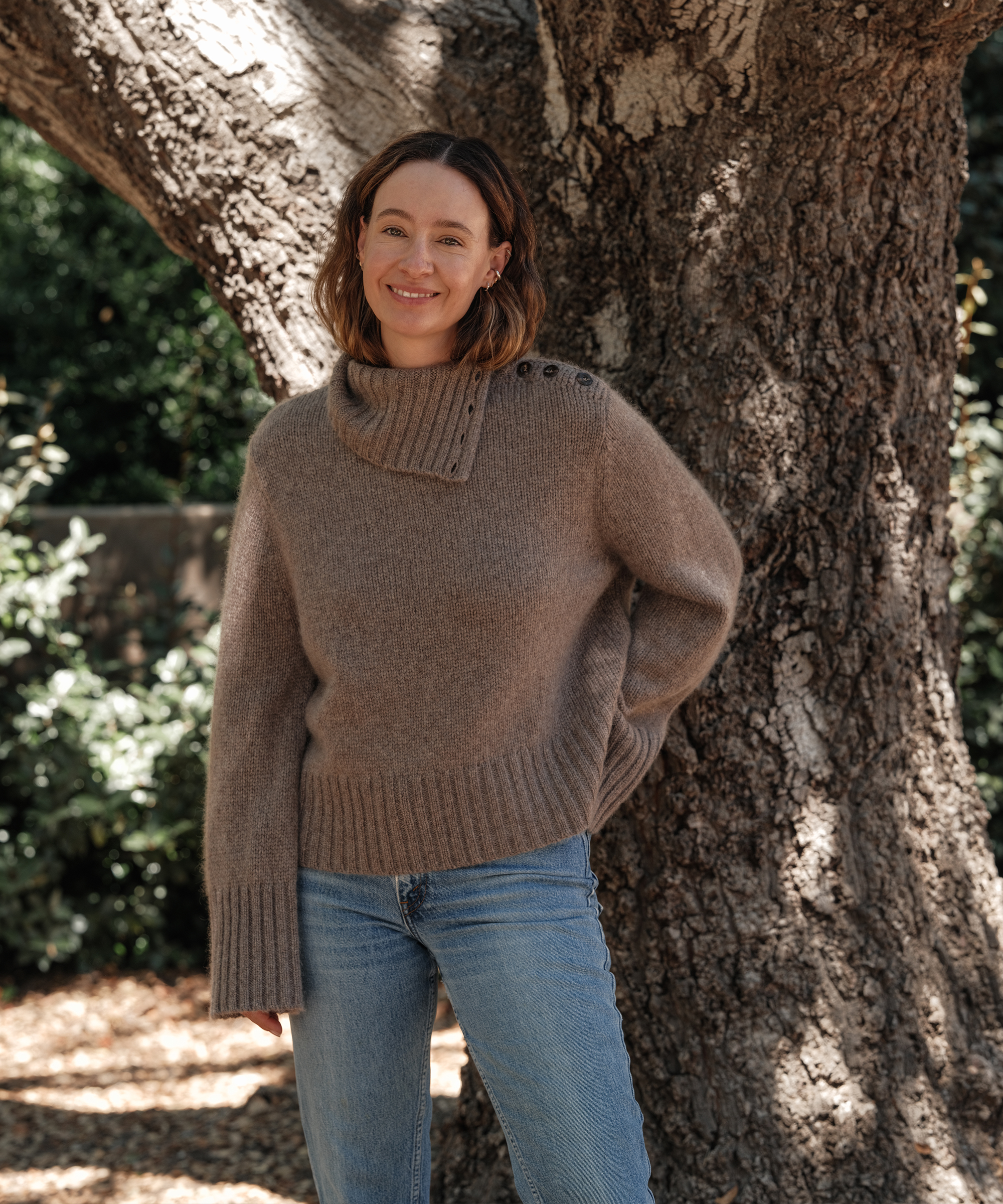 jenni in a taupe cashmere sweater and denim standing in front of a tree