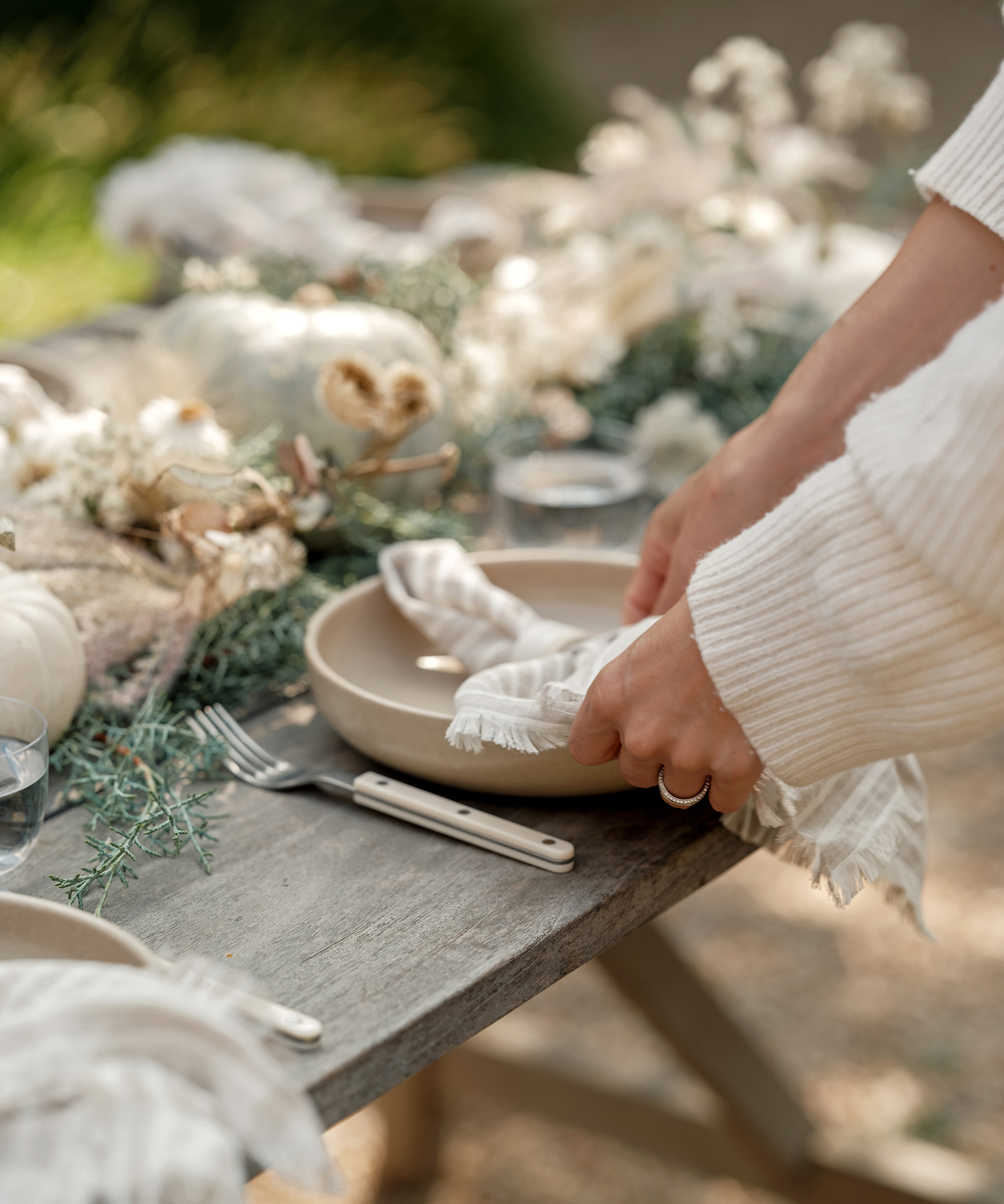 a hand placing a napkin on a taupe plate on a wooden outdoor table