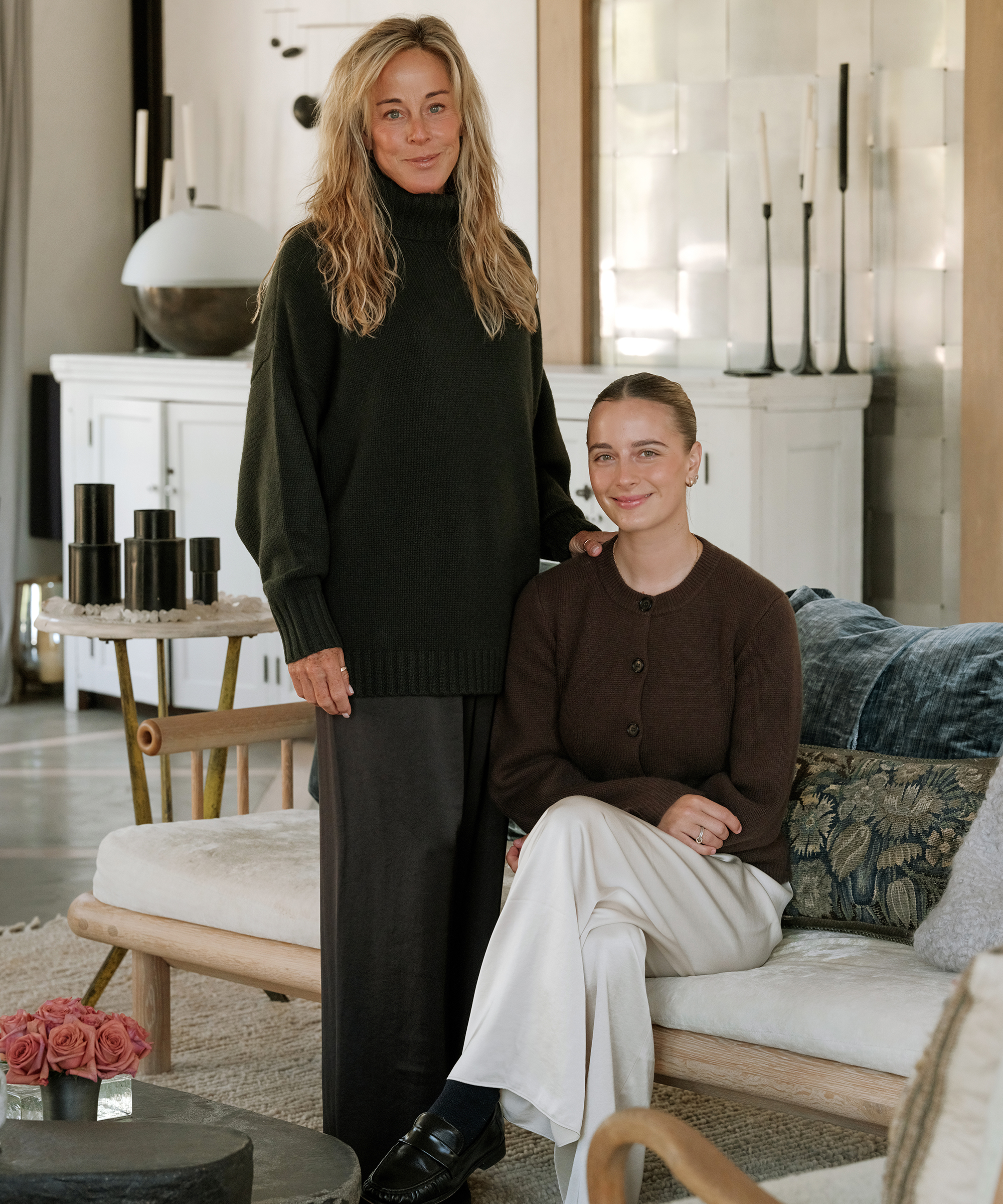 a woman standing next to her daughter sitting on a couch in a brown cardigan and ivory pants
