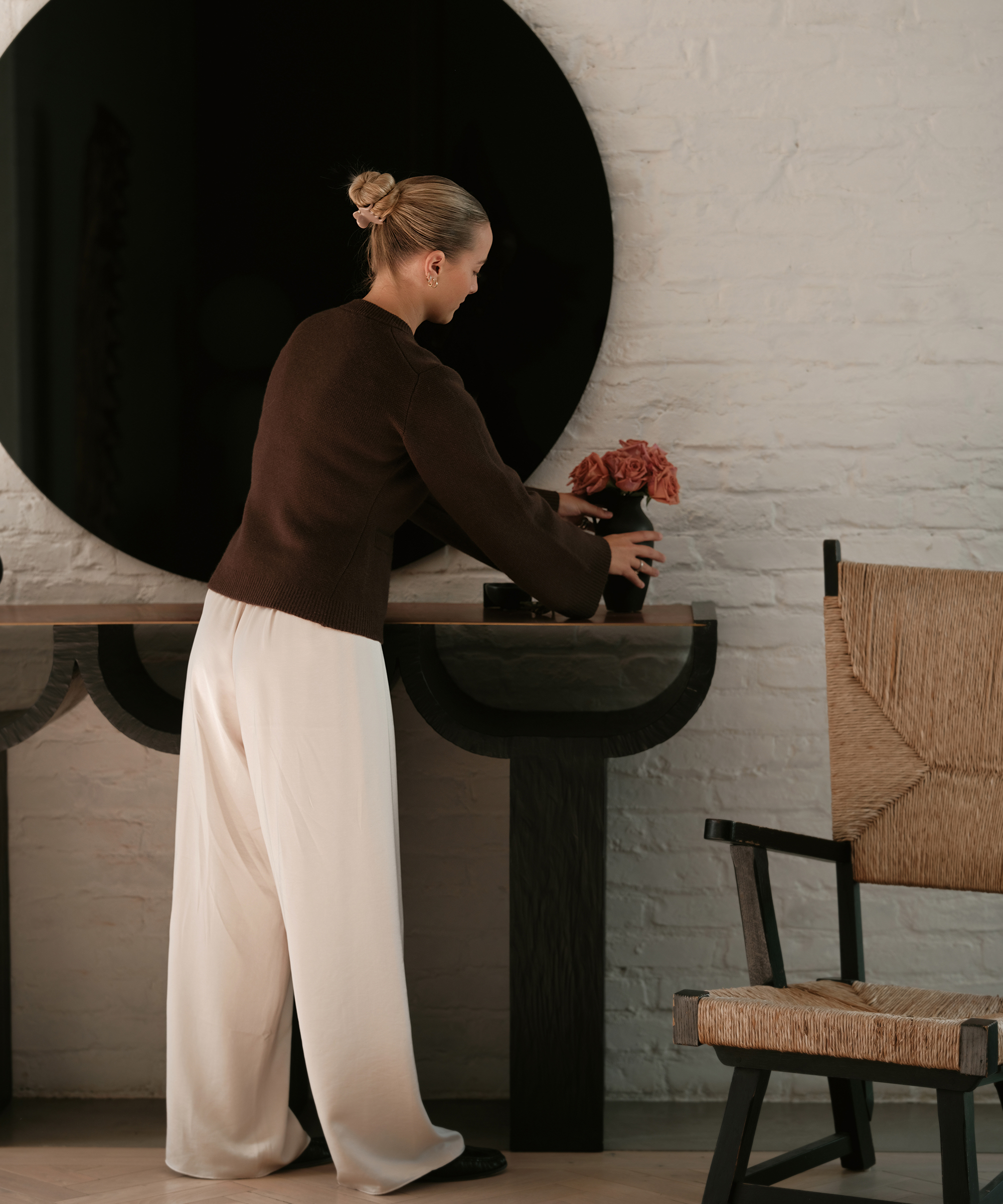 a woman placing a vase on a black wooden sideboard wearing ivory silk trousers and a brown cardigan