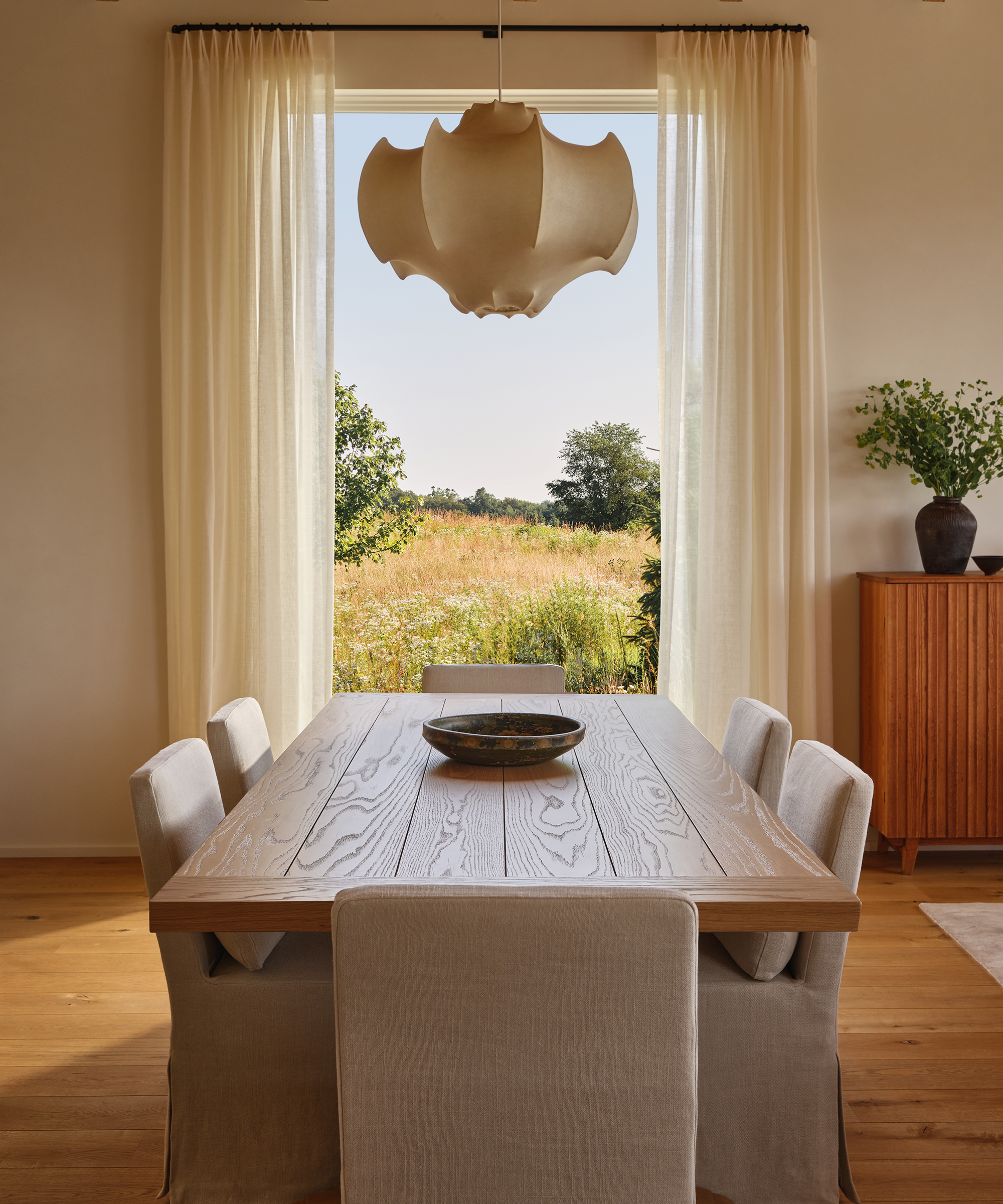 Step into this inviting dining room, often featured in home tour highlights, with a wooden table, six cream chairs, sheer curtains, and a large window framing views of green fields and trees. A stylish sideboard with vases sits to the right.