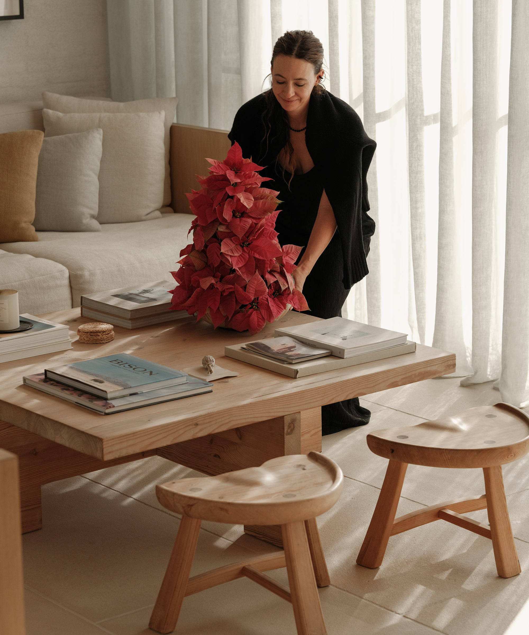 A woman in holiday dressing places a poinsettia plant on a wooden coffee table in a cozy, sunlit living room with neutral decor, books, and wooden stools.