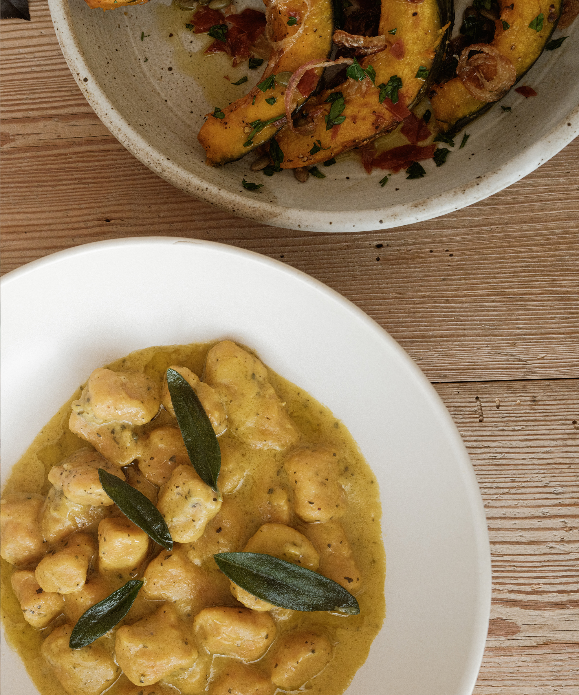 A white plate of gnocchi in creamy yellow sauce, garnished with fresh sage, sits beside a bowl of roasted pumpkin with herbs and crispy onions—a perfect holiday dinner spread on a rustic wooden table.