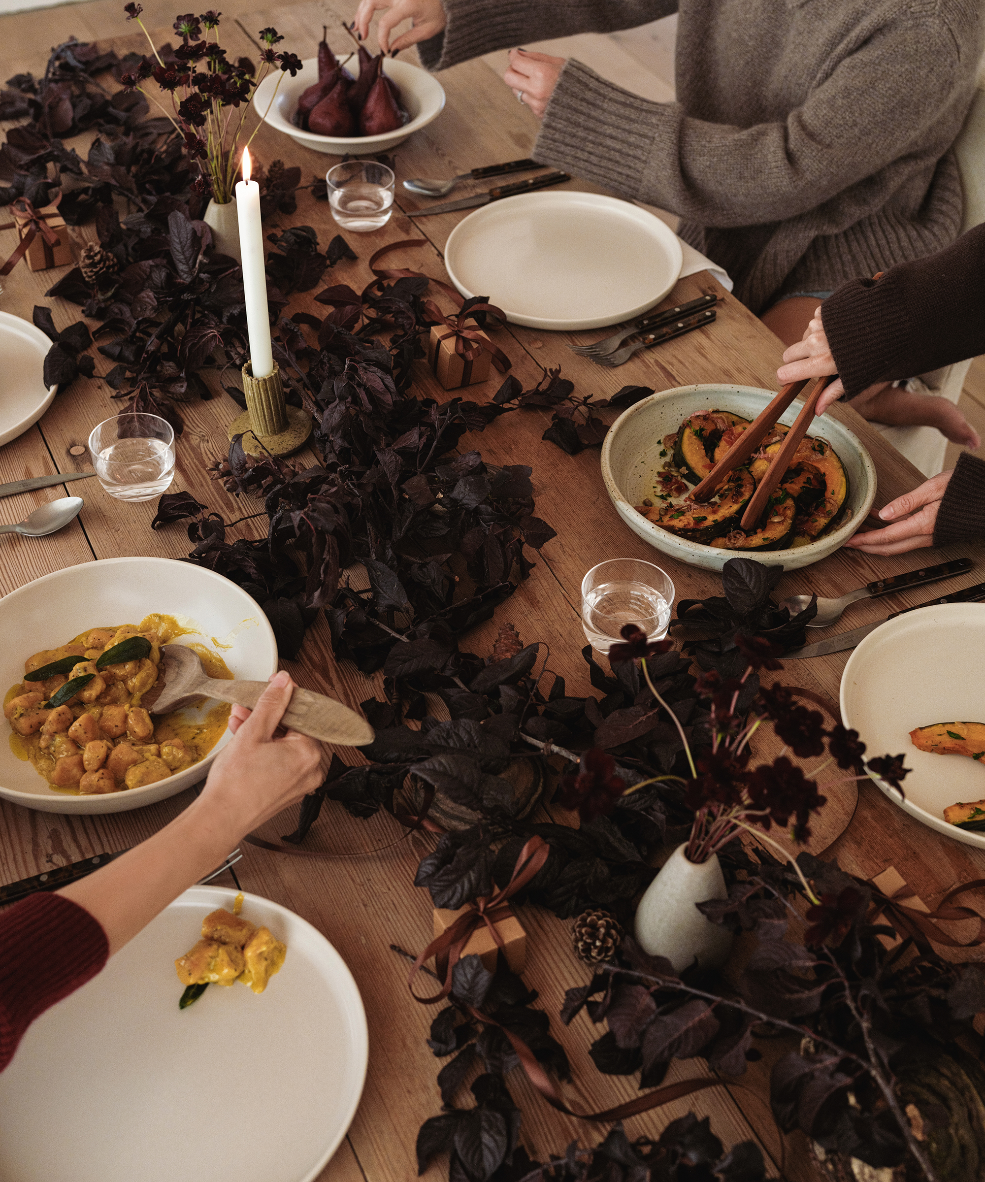 Three people sit around a wooden table set for a holiday dinner, with plates of food, glasses, cutlery, a lit candle, and dark purple floral arrangements, serving and sharing dishes in a cozy, intimate setting.