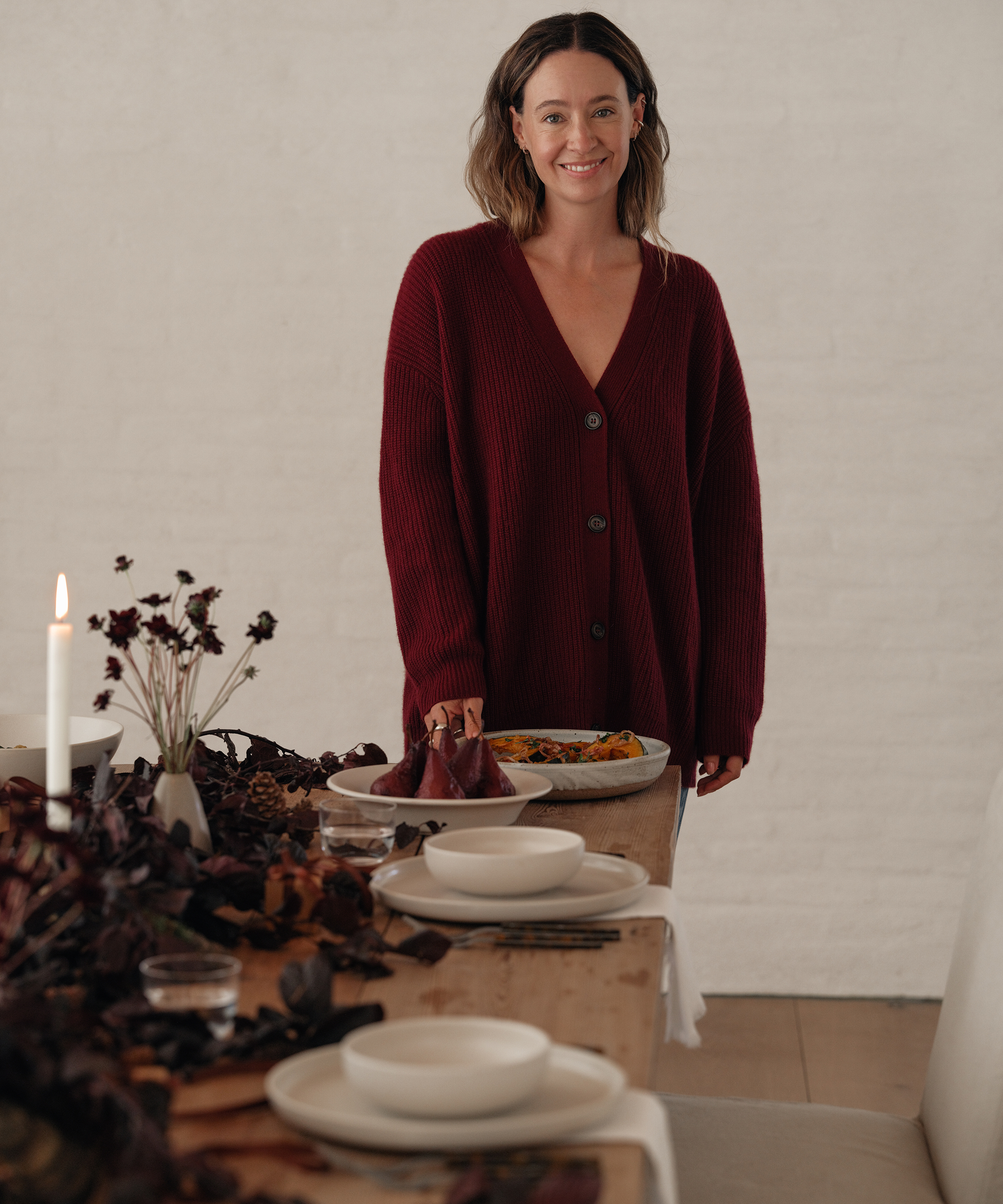 A woman in a maroon cardigan stands at a decorated dining table set for a holiday dinner, with candles, dark flowers, and plates, smiling at the camera while holding a dish of food.