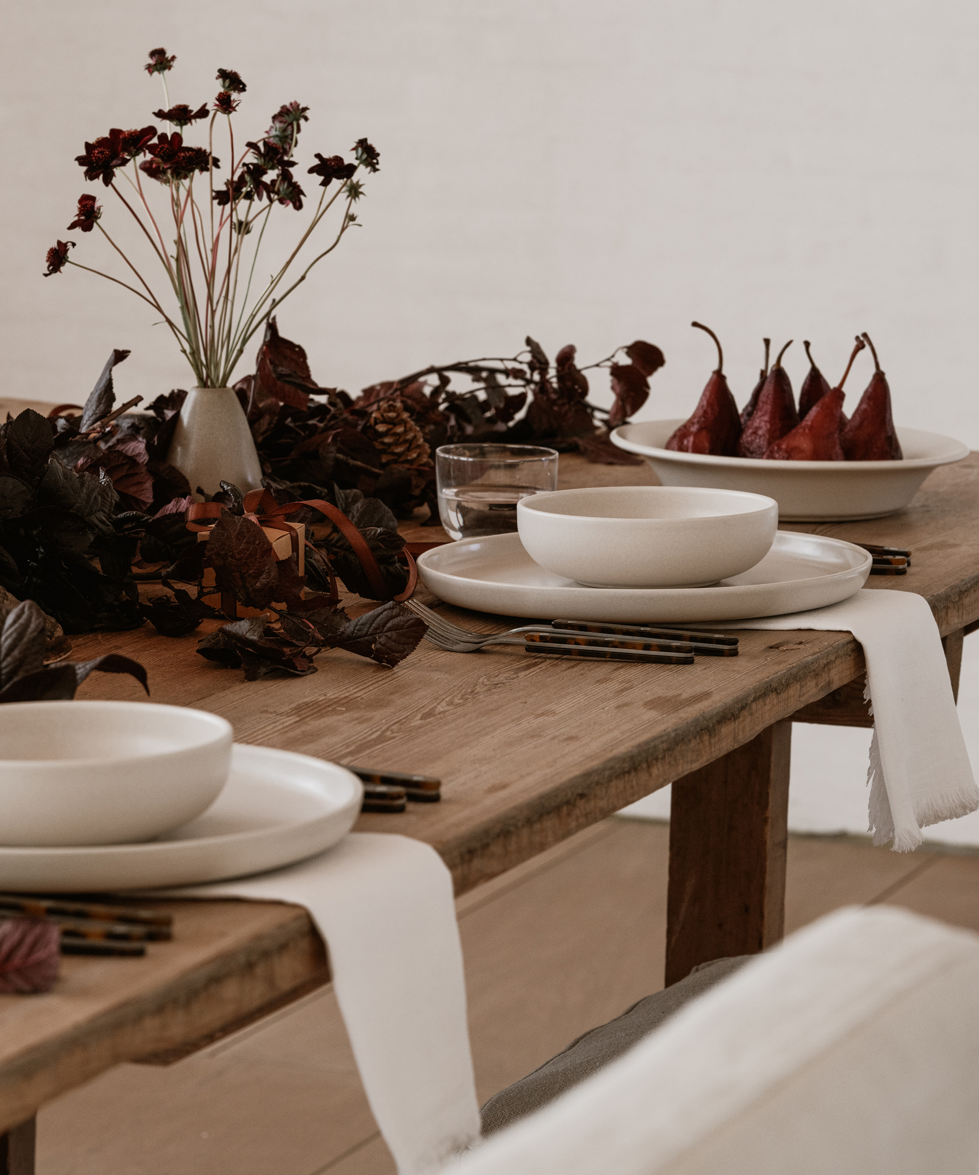 A rustic wooden table is set for a holiday dinner with white plates and bowls, black-handled cutlery, white napkins, a vase of dark red flowers, autumn leaves, pinecones, and a plate of poached pears in the background.