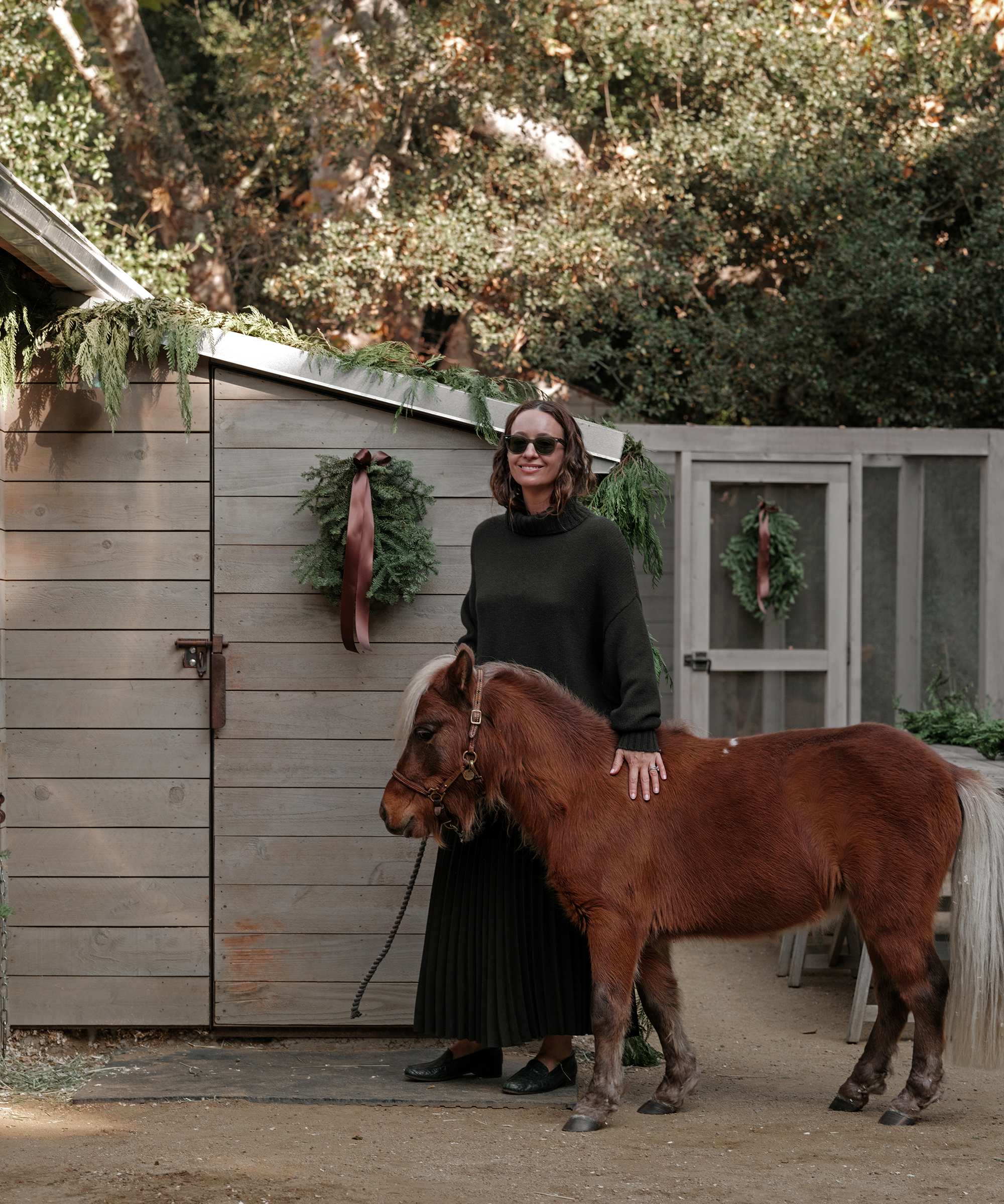 jenni wearing a pleated skirt and a cashmere turtleneck in forest green standing next to her stable with a mini horse